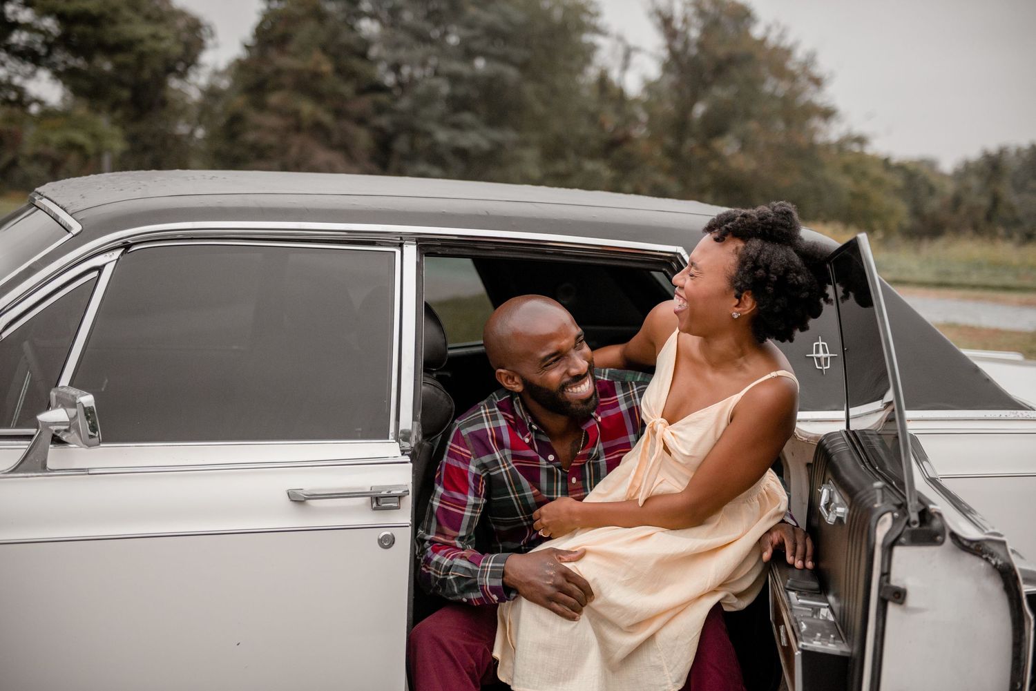 A joyful couple laughing together inside a classic white vintage car during an engagement photo session.