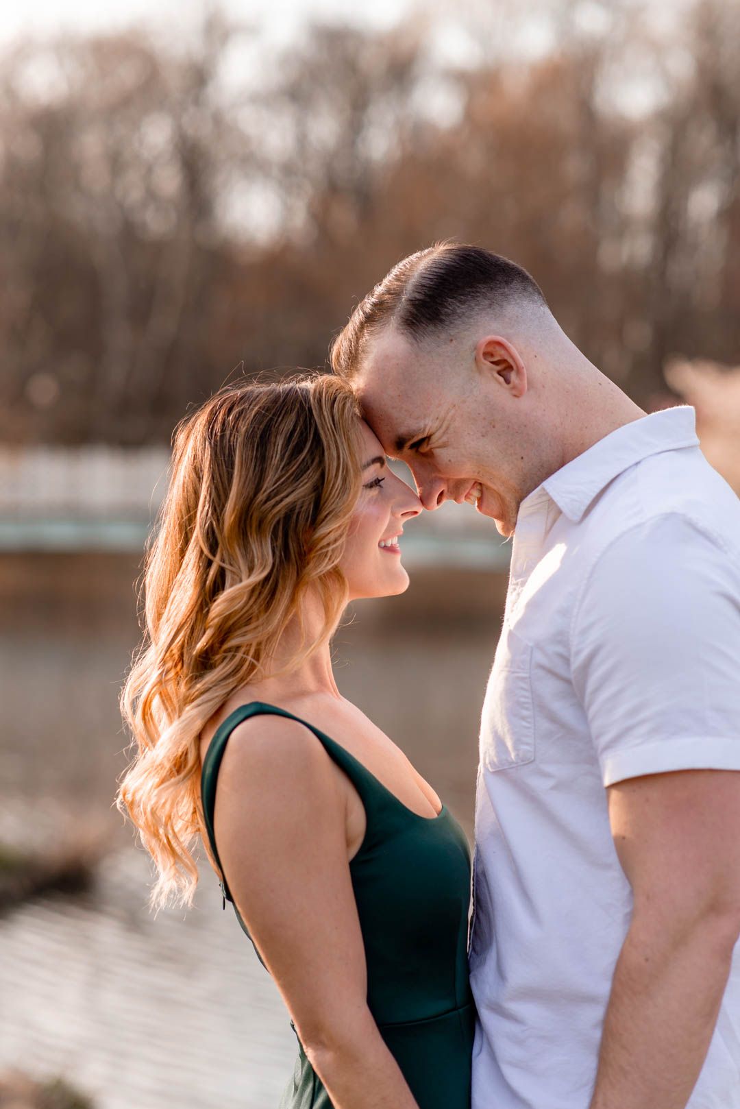 Couple touching foreheads and smiling intimately near a sunlit lake during golden hour engagement session.