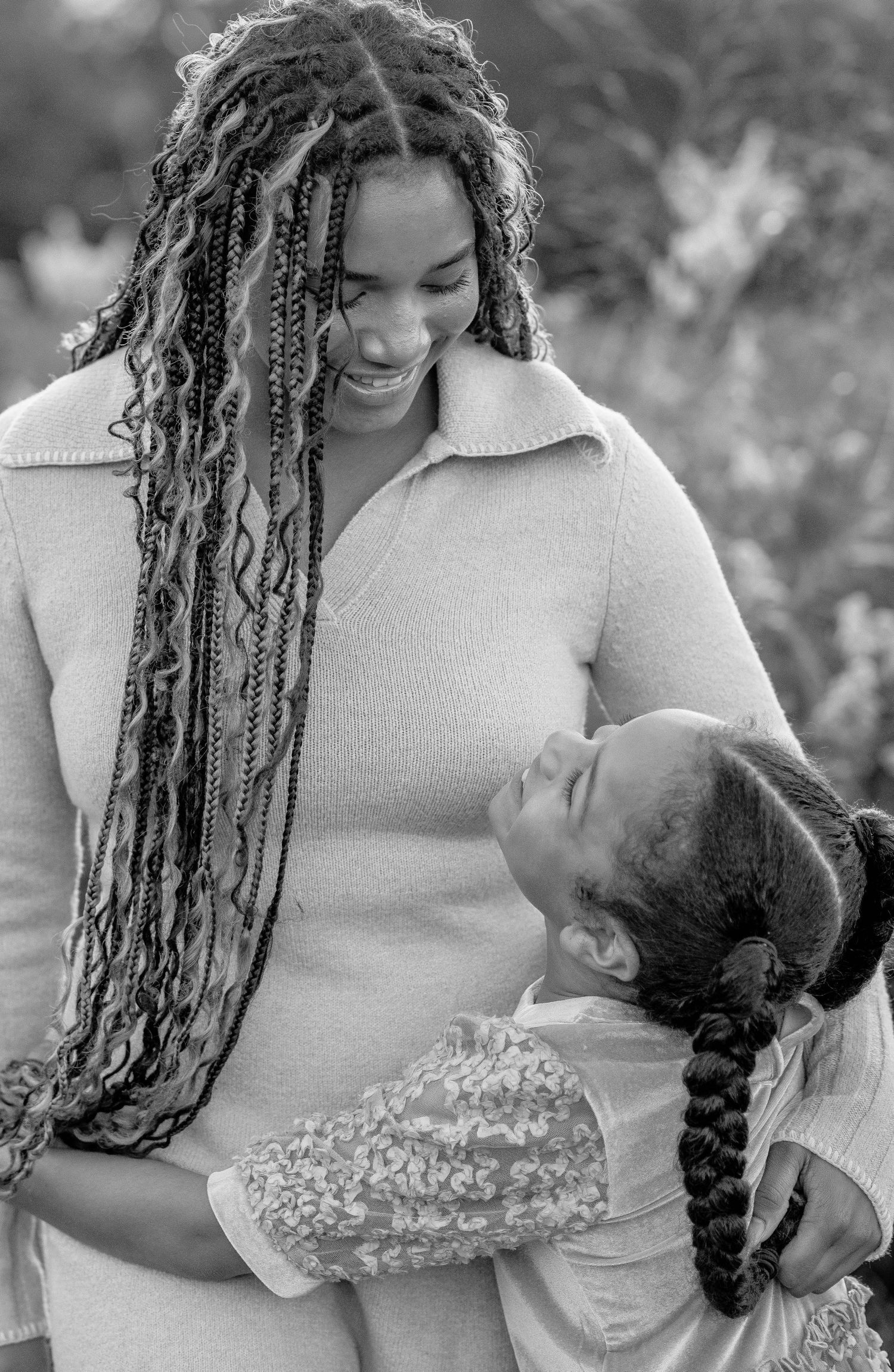 Black and white photo of a mother with long braids smiling at her young daughter in an outdoor setting.