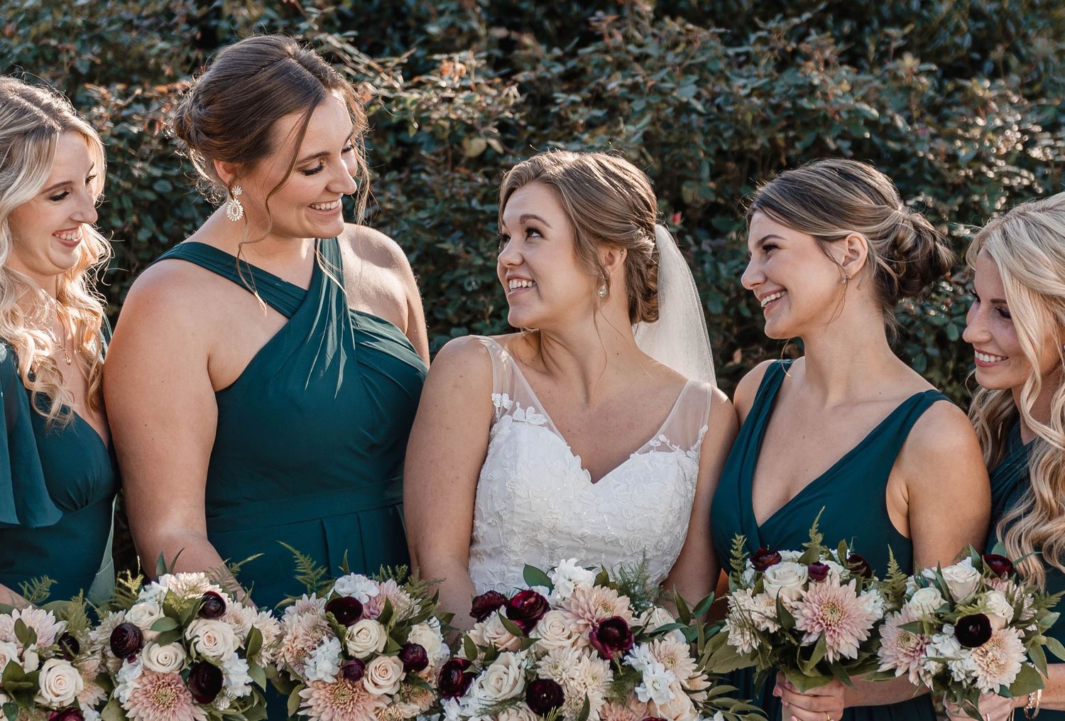 Bride smiling with four bridesmaids in dark green gowns holding blush and burgundy bouquets outdoors.