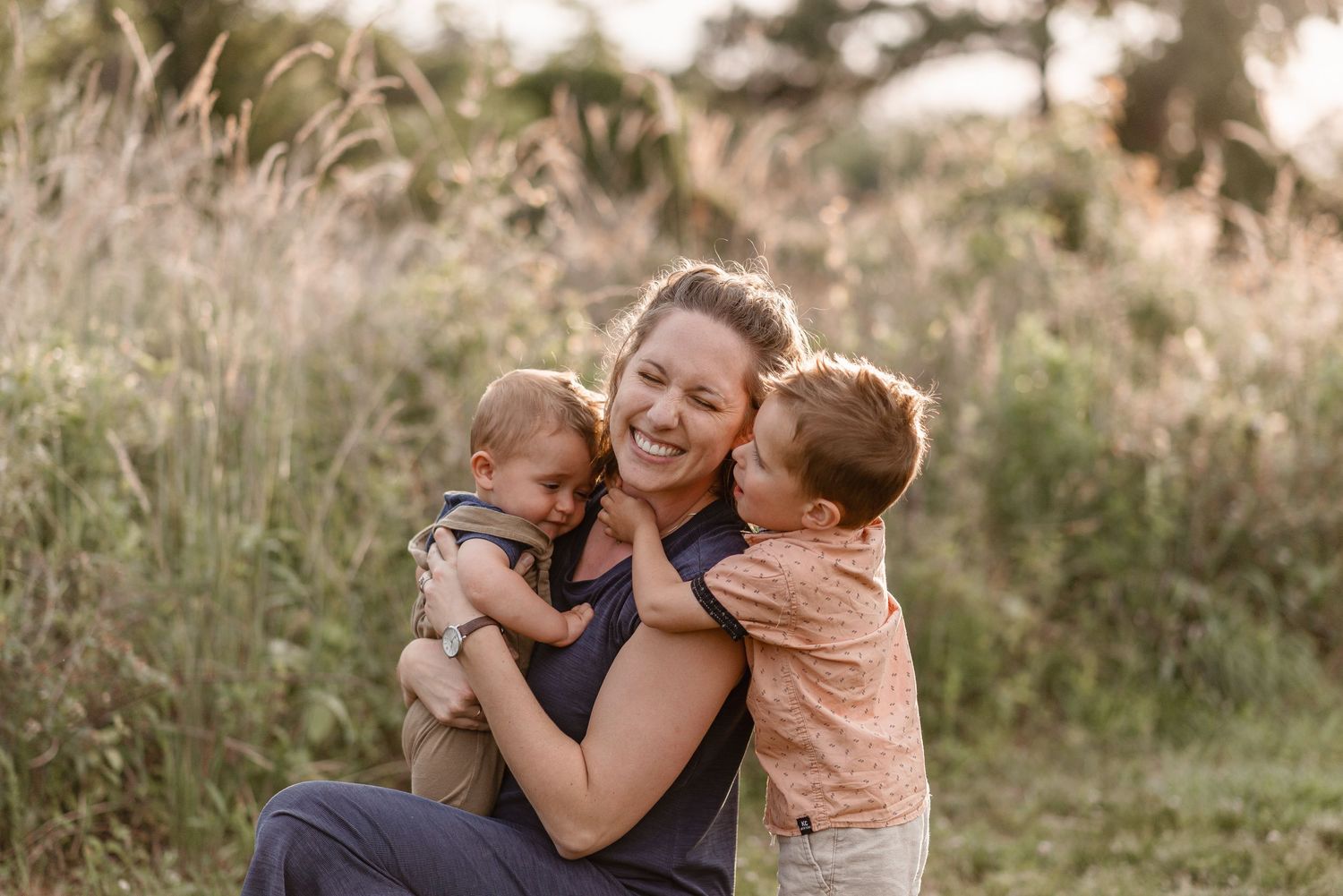 A smiling mother sits in a meadow holding her baby while her young son gives her a sweet kiss on the cheek.