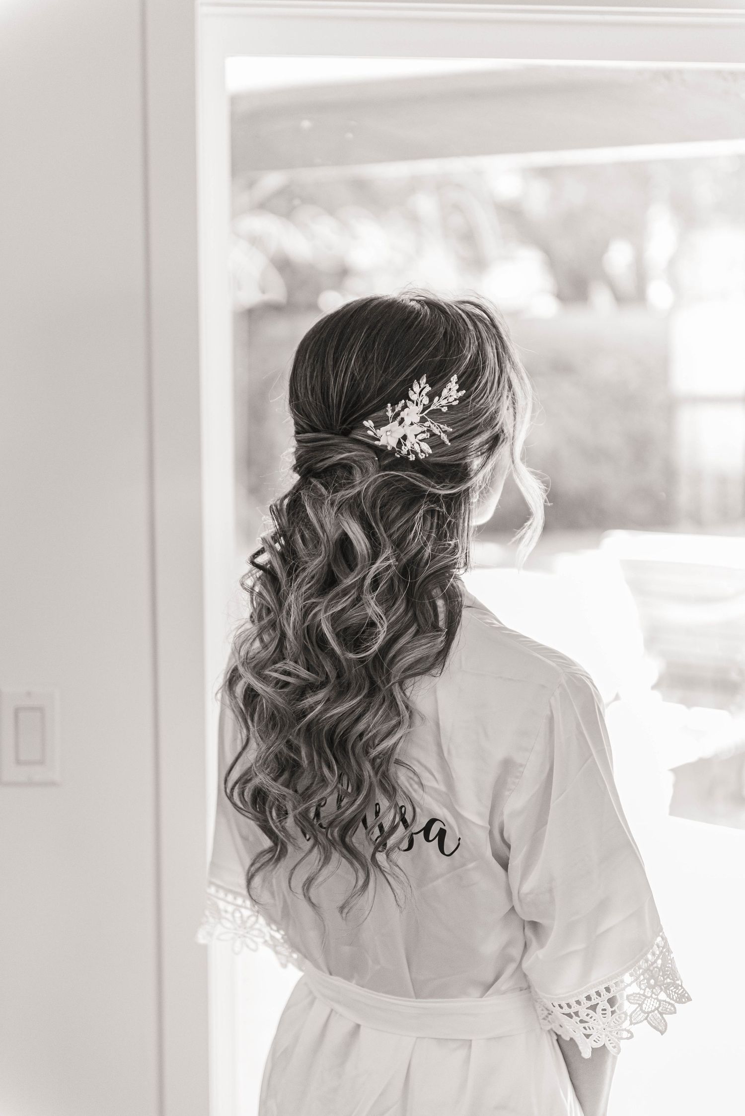 Black and white photo of bride in personalized robe with elegant half-up curled hairstyle and hair accessory.