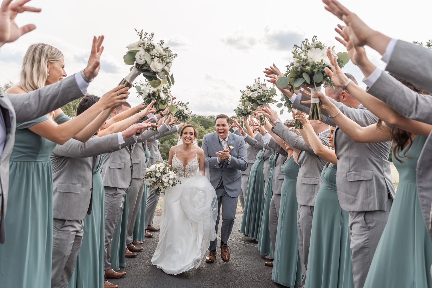 Bride and groom walk through tunnel of raised bouquets held by wedding party in sage and gray.
