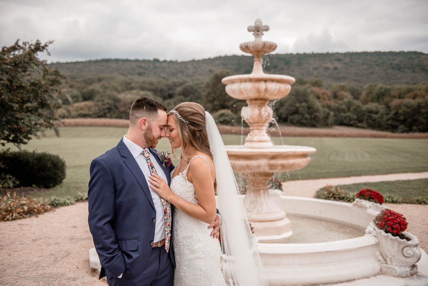 Bride and groom touch foreheads romantically in front of stone fountain with mountain backdrop.