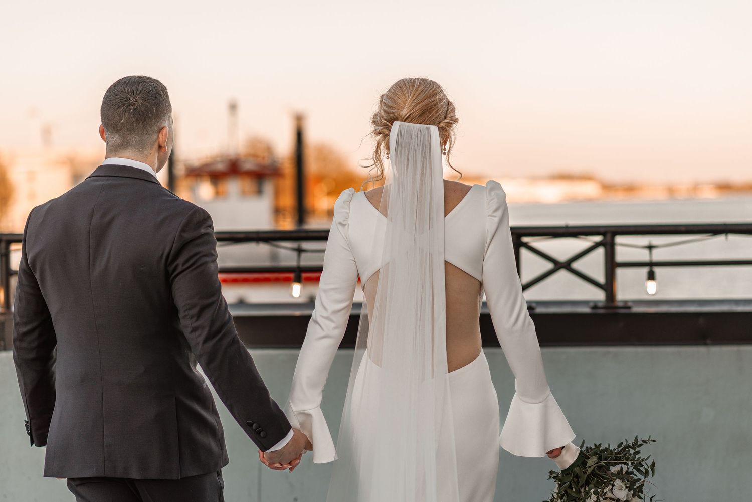 Bride in backless long-sleeve gown holding hands with groom overlooking waterfront at sunset.