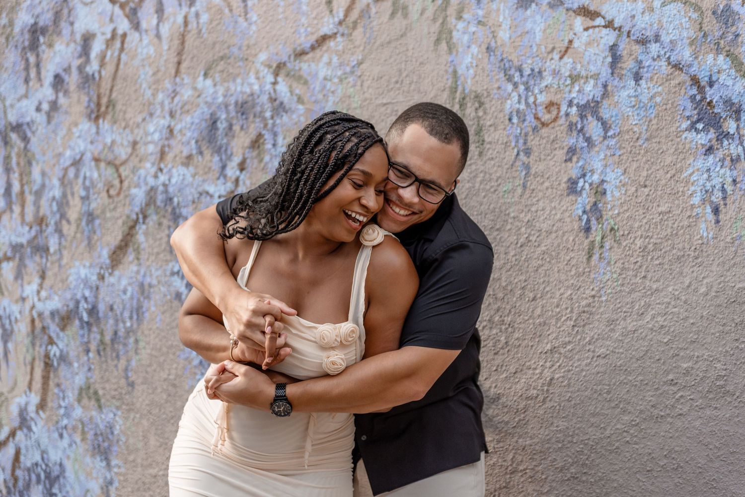 Happy couple embracing and laughing in front of a blue floral wisteria wall mural.