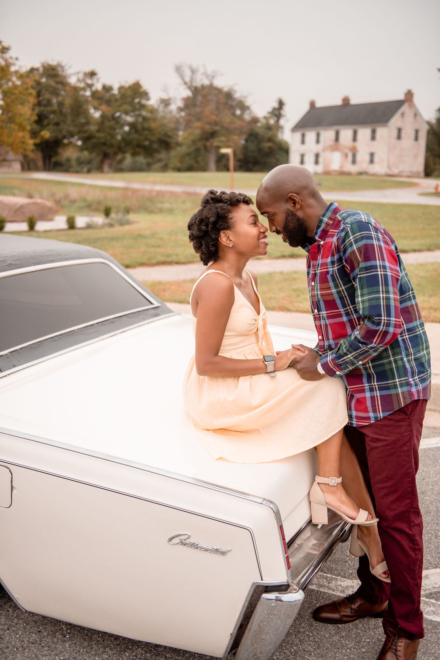 Romantic couple sharing intimate moment leaning against classic white Chevrolet car near historic farmhouse.