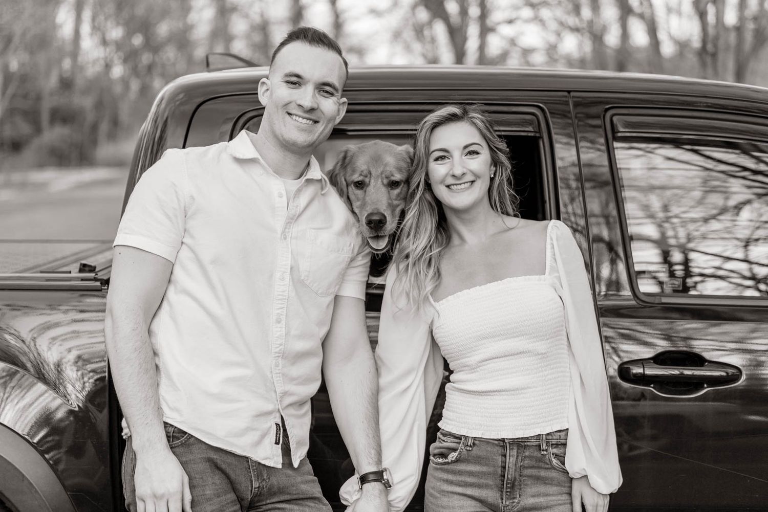 Black and white photo of smiling couple with golden retriever dog leaning against vintage truck outdoors.