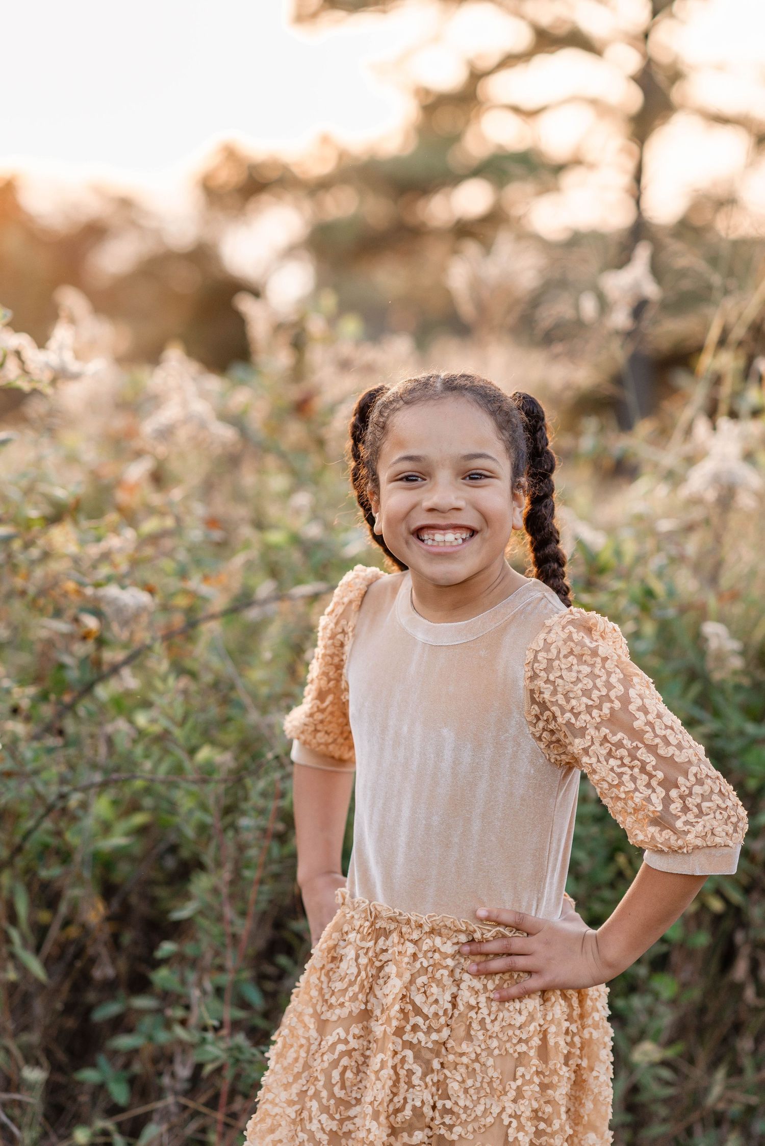 A young girl in a gold textured dress smiles brightly with hands on hips in a sunny outdoor setting.