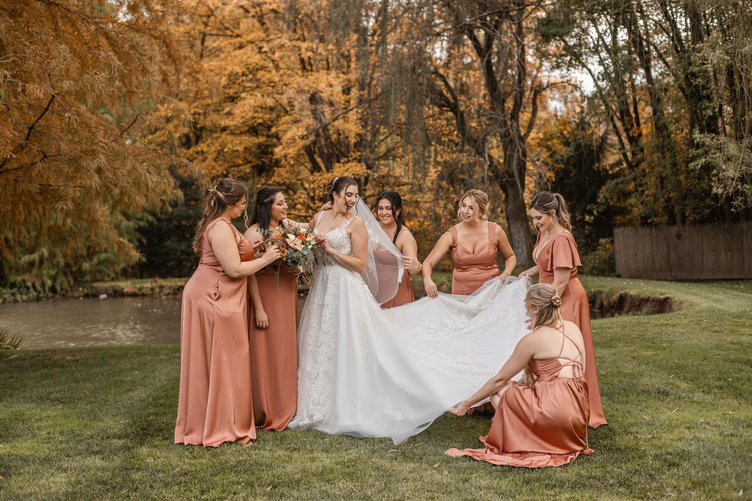Bride in white lace gown surrounded by bridesmaids in rust-colored dresses among autumn foliage.