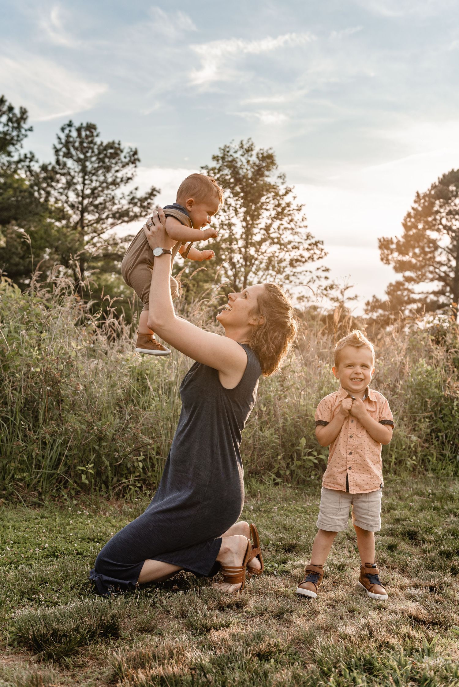 A mother kneels on grass lifting her baby in the air while her toddler son smiles beside her at golden hour.