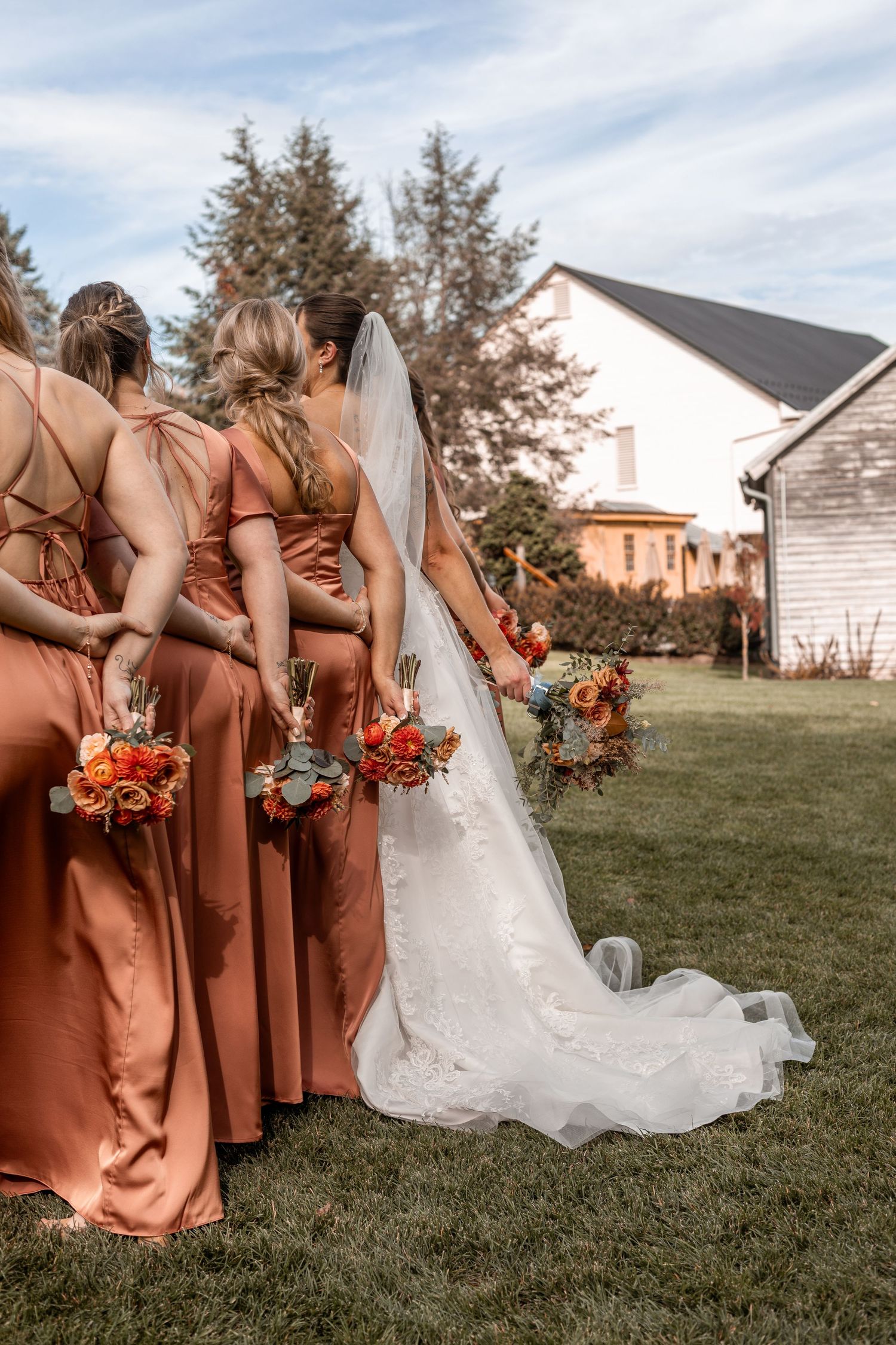 Bride in lace gown with long veil stands with bridesmaids in terracotta satin dresses on green lawn.