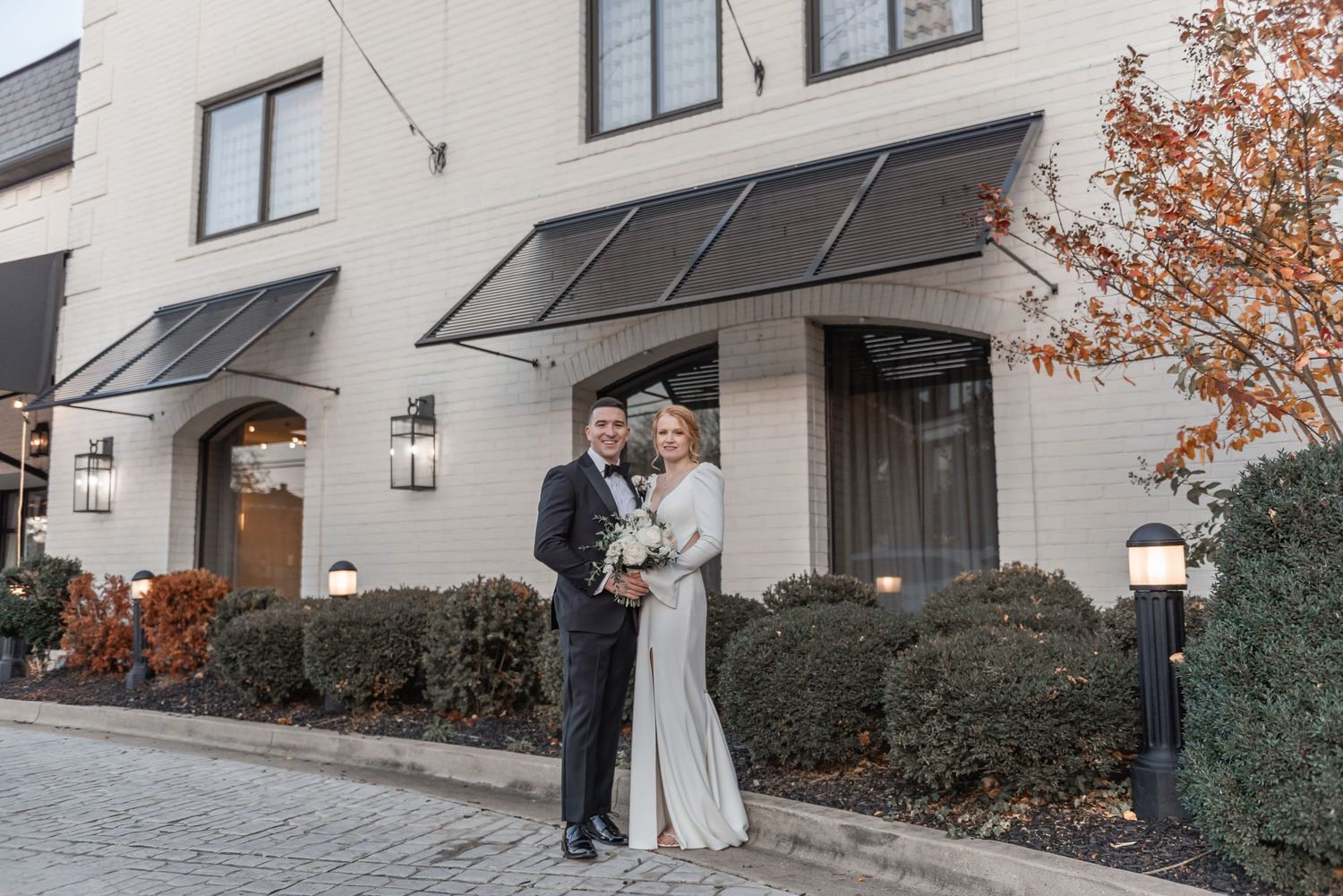Bride and groom pose together outside an elegant white brick building with autumn foliage.