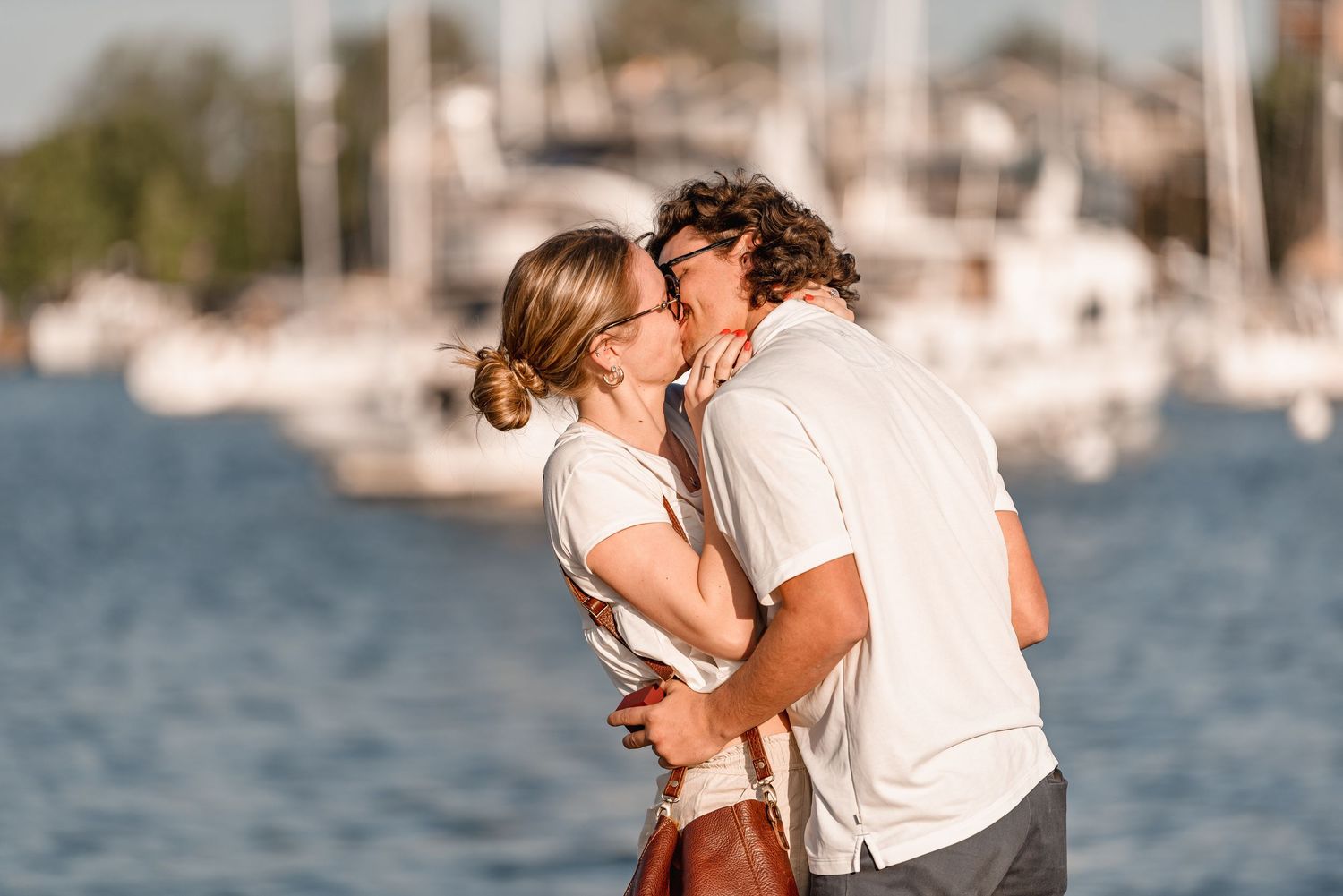 Newly engaged couple sharing a romantic kiss on a sunny marina dock with sailboats in background.