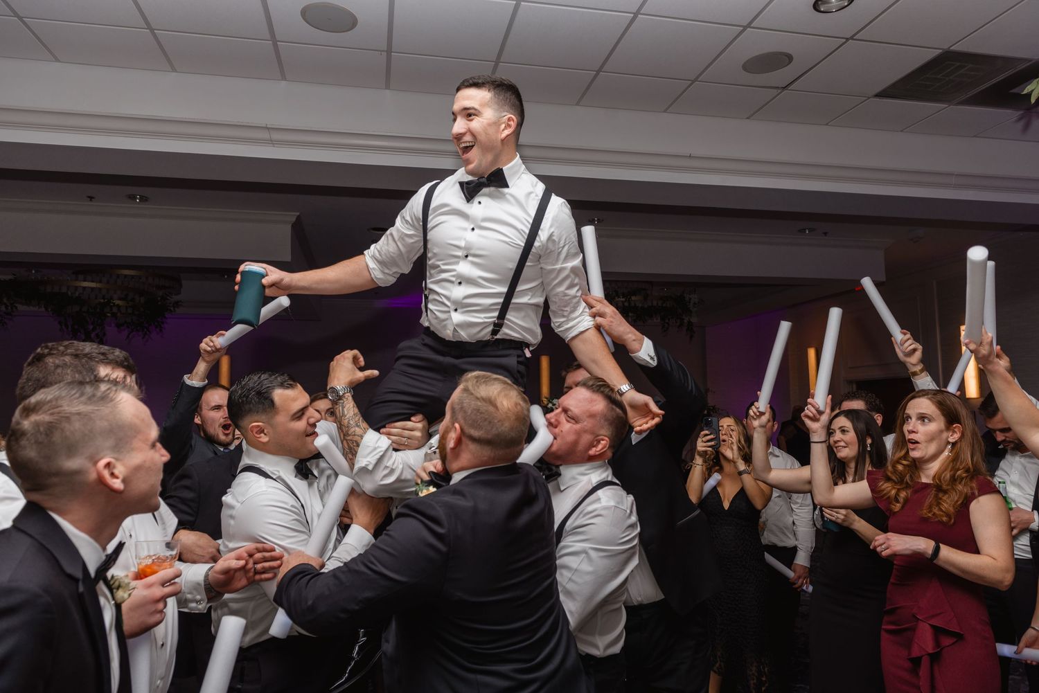 Groom lifted by groomsmen on packed dance floor during lively wedding reception celebration with light sticks.