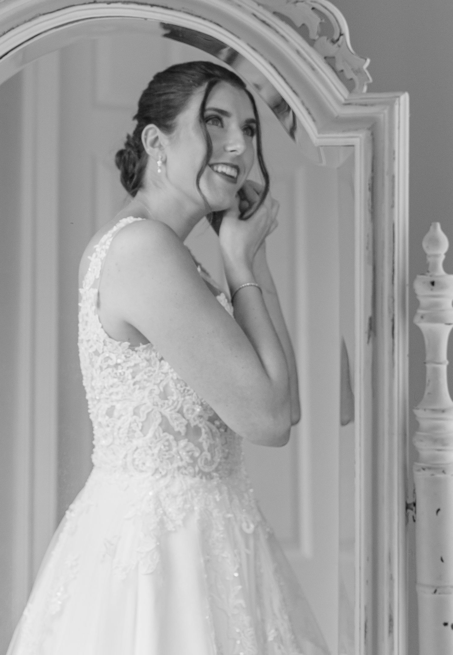 Joyful bride in a lace wedding gown laughs beside an ornate vintage mirror in a black and white photo.