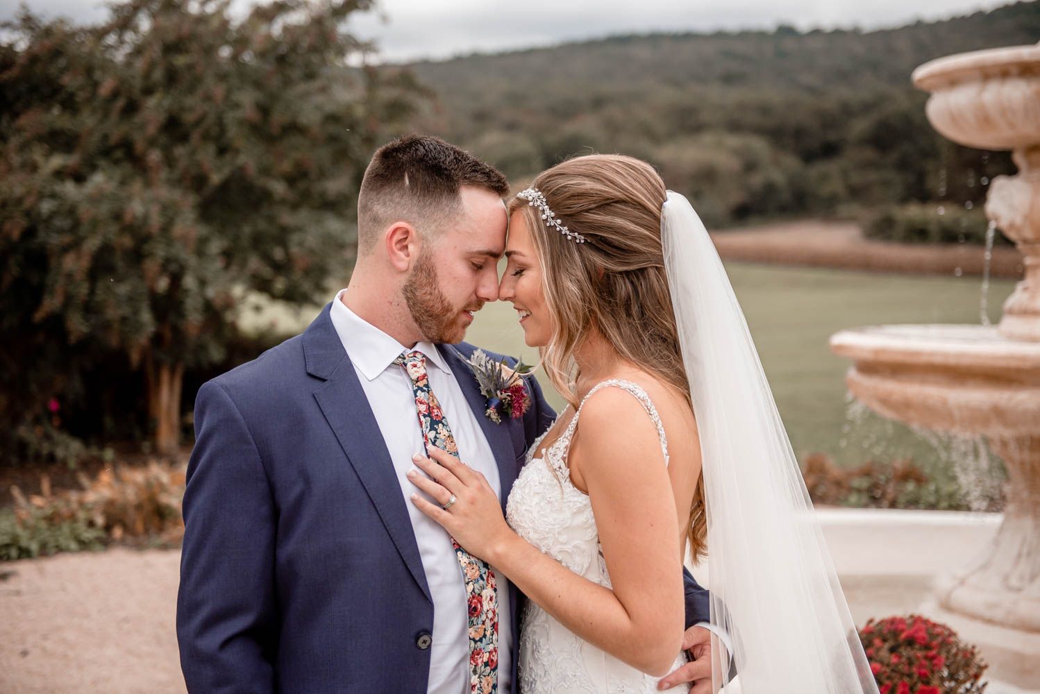 Bride and groom touching foreheads tenderly near stone fountain with lush green hills behind them.