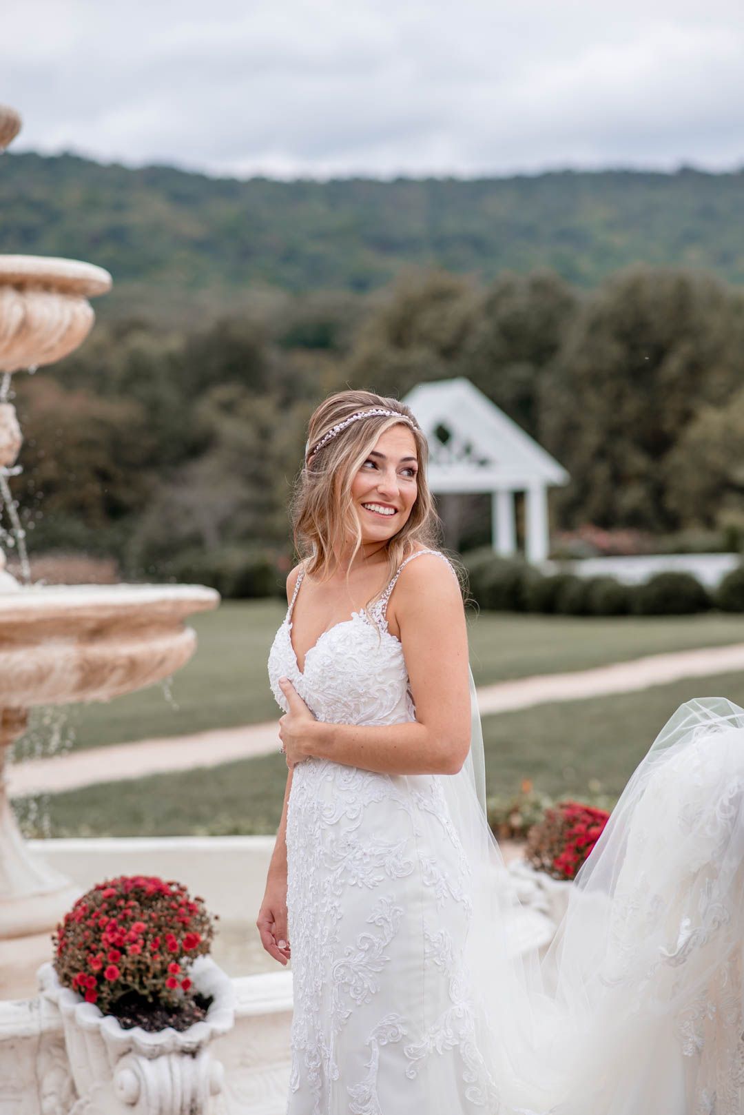 Smiling bride in a lace gown poses near a stone fountain with a mountain and white chapel in the background.