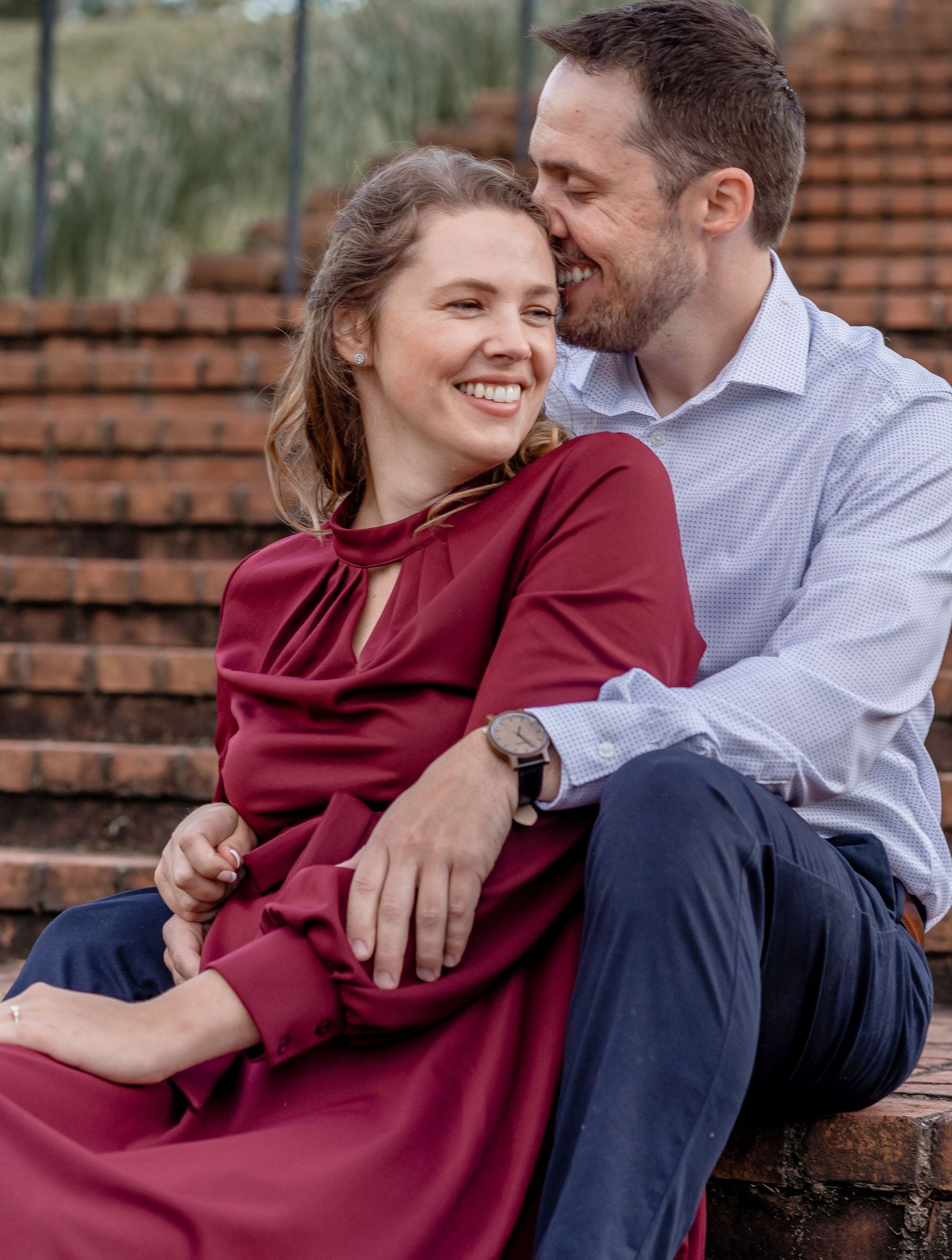 Couple sitting on brick steps, woman in burgundy dress laughing as man whispers in her ear.