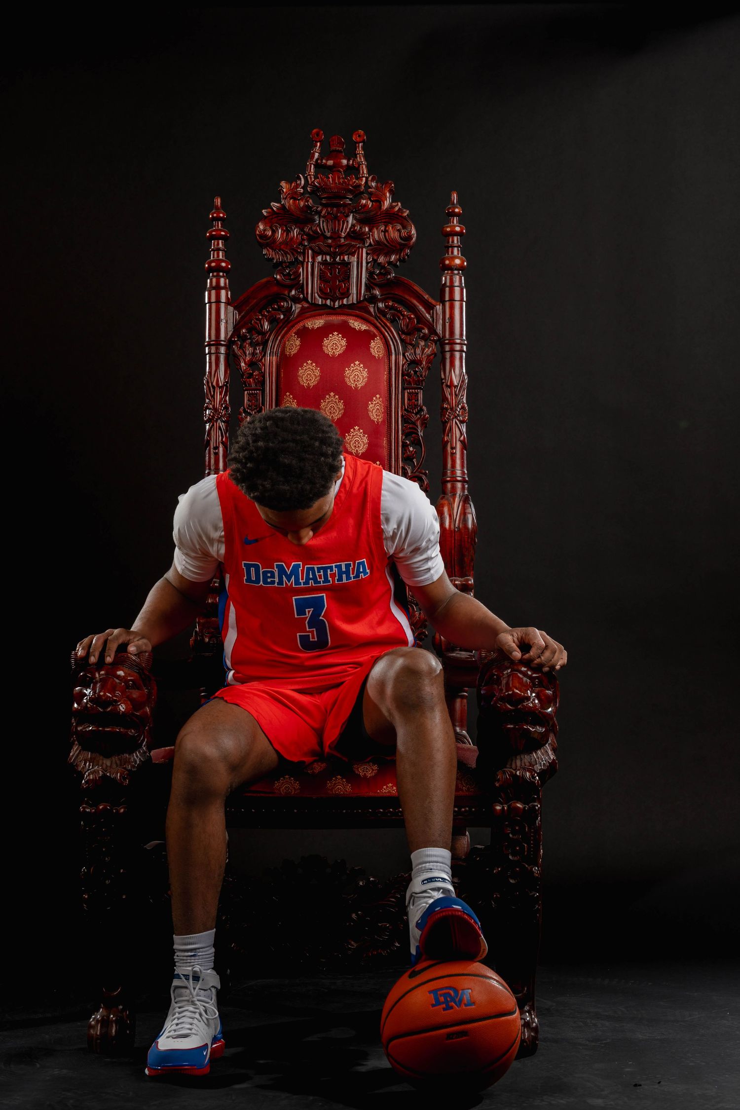 Basketball player #3 in DeMatha jersey sits on ornate red throne with ball.