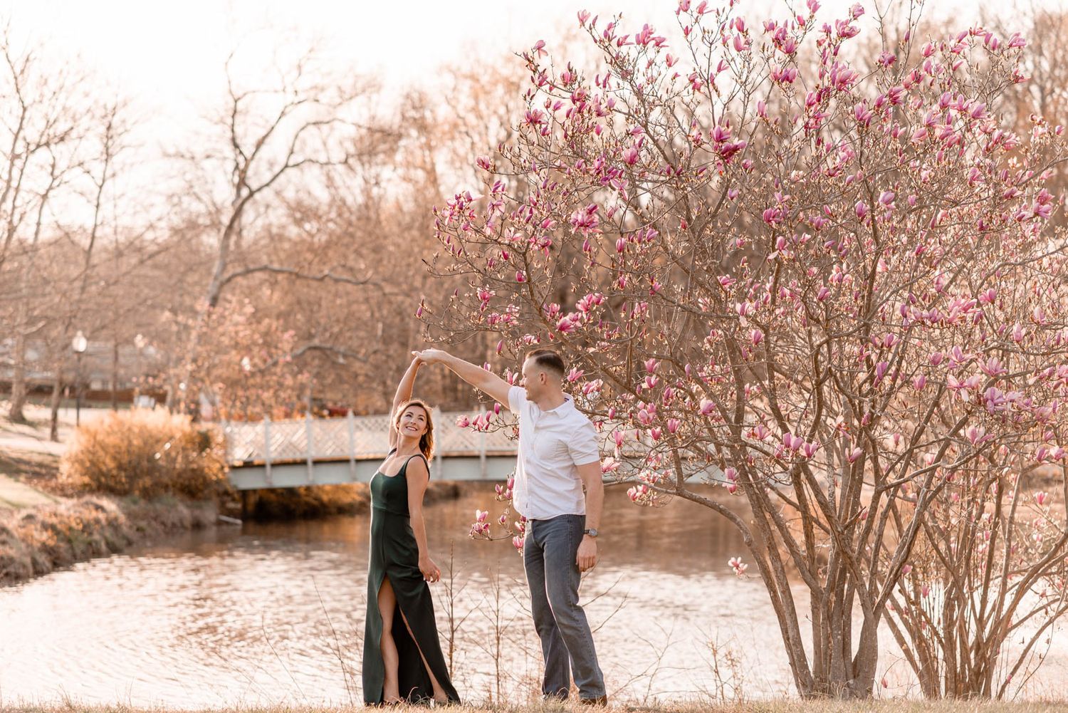 Couple dancing together near a pond with blooming pink magnolia trees during golden hour engagement photos.