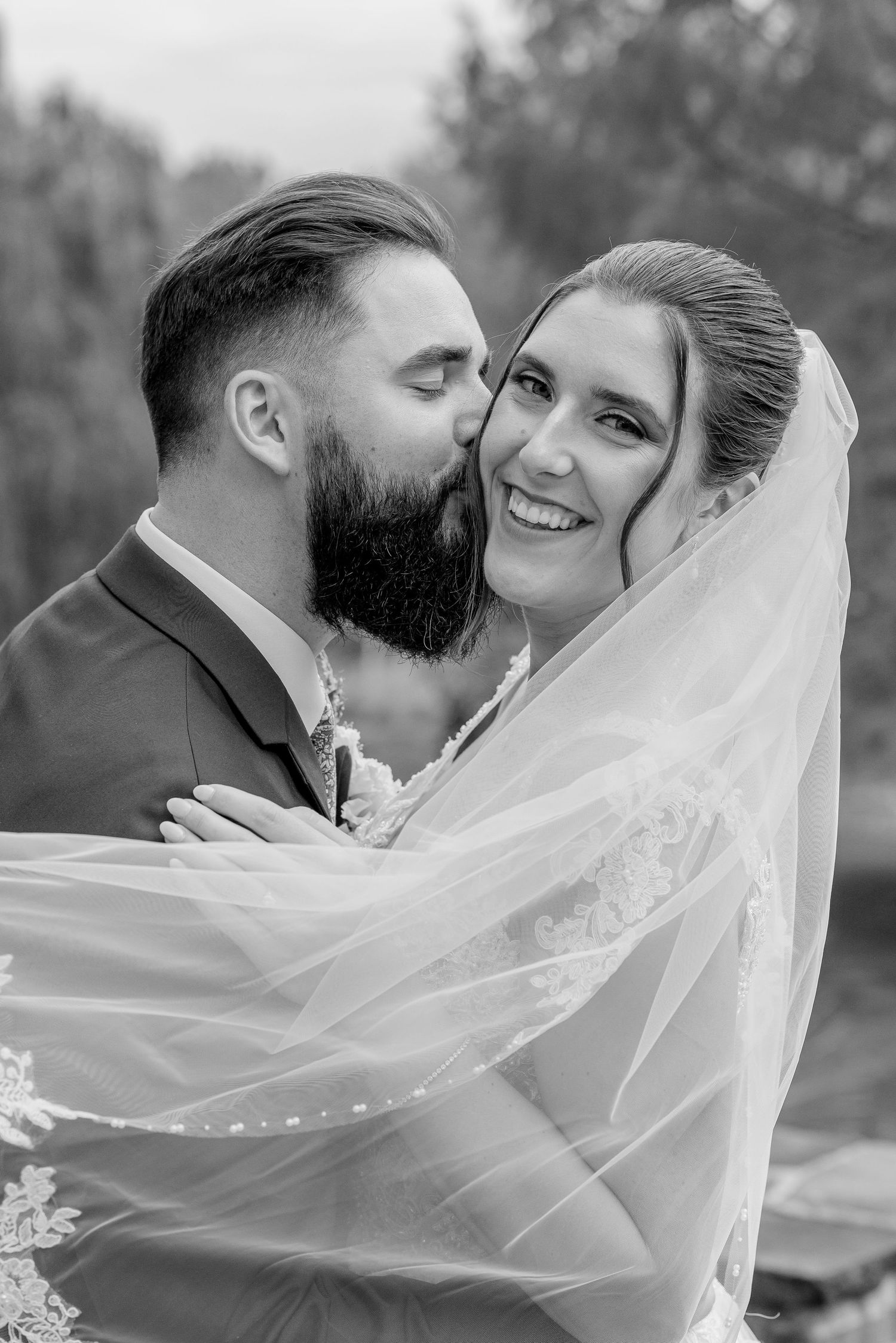 Black and white photo of a bearded groom kissing his smiling bride on the cheek under her veil.