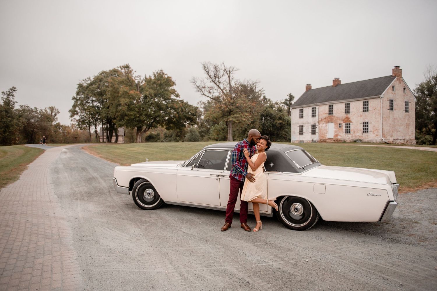 A couple shares a romantic kiss leaning against a classic white vintage car on a scenic country road.
