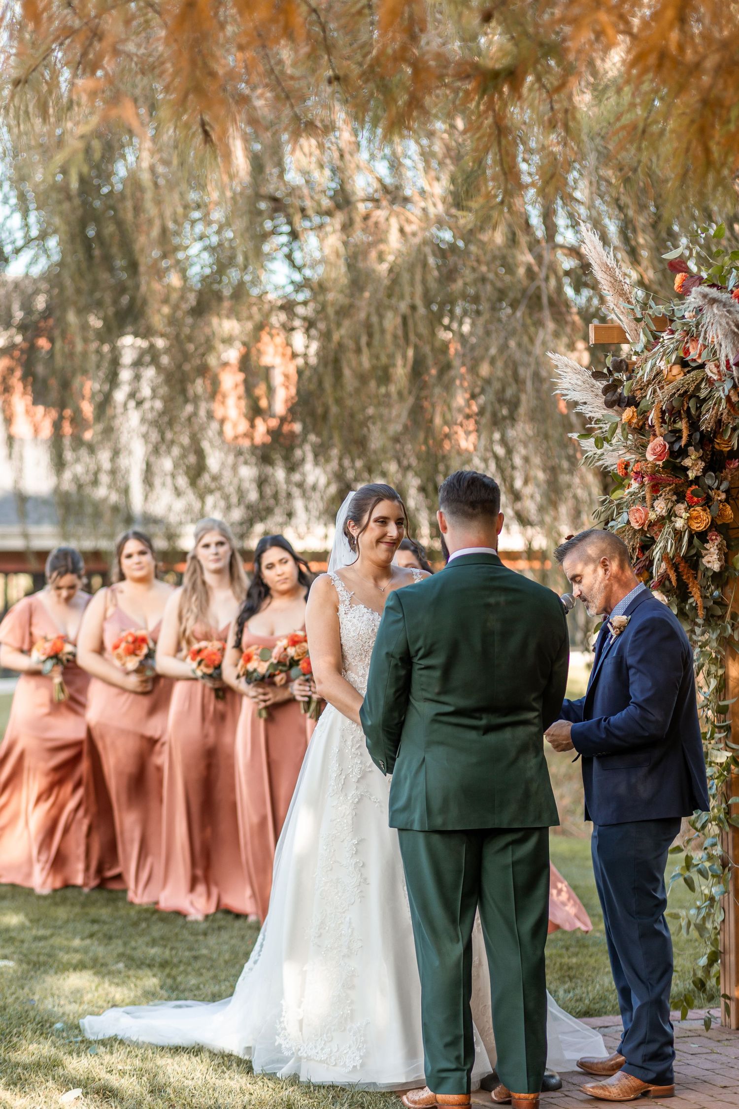 Bride and groom exchange vows outdoors with bridesmaids in dusty rose dresses standing beneath a weeping willow tree.
