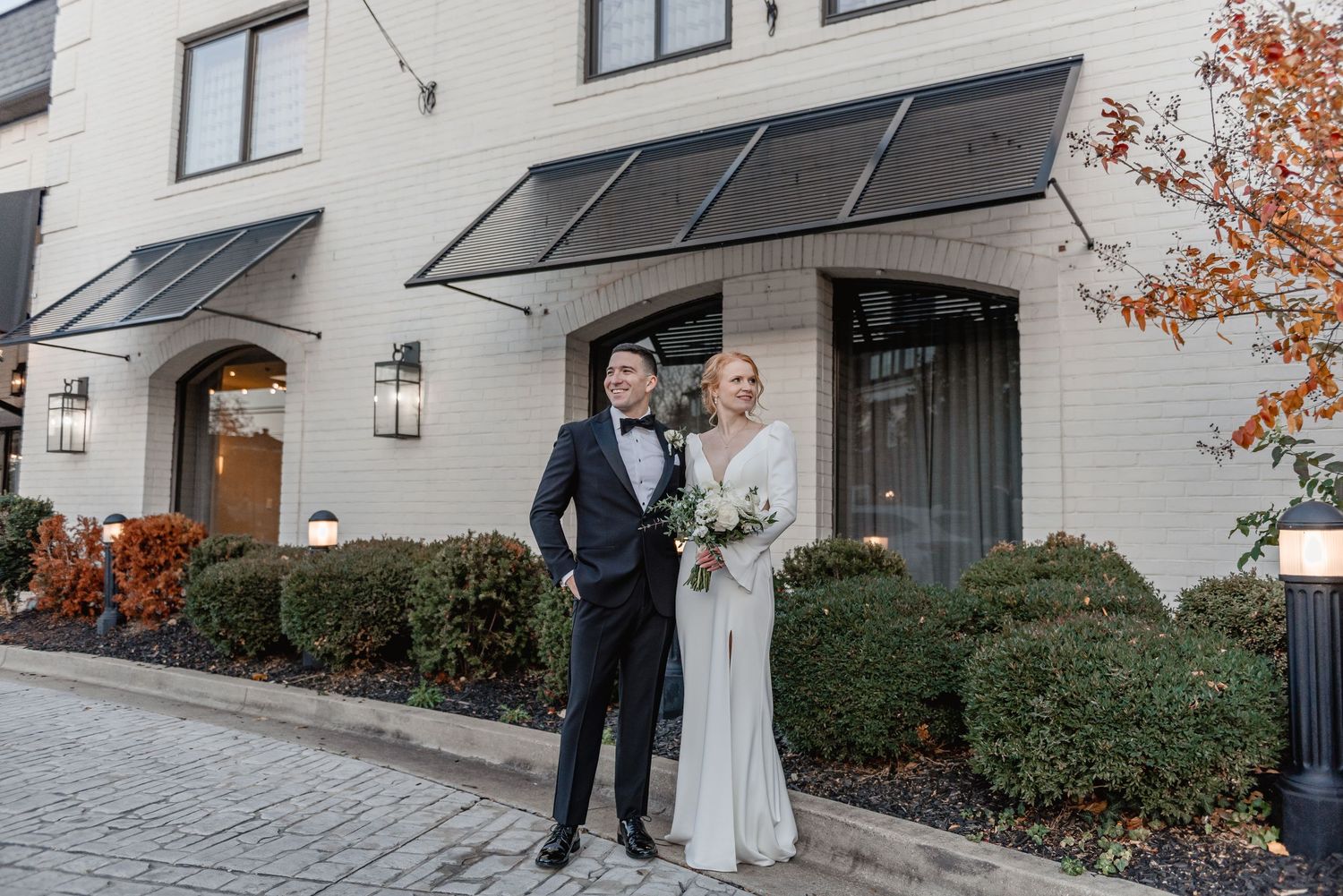 Bride and groom pose together outside a modern white brick building during their fall wedding.