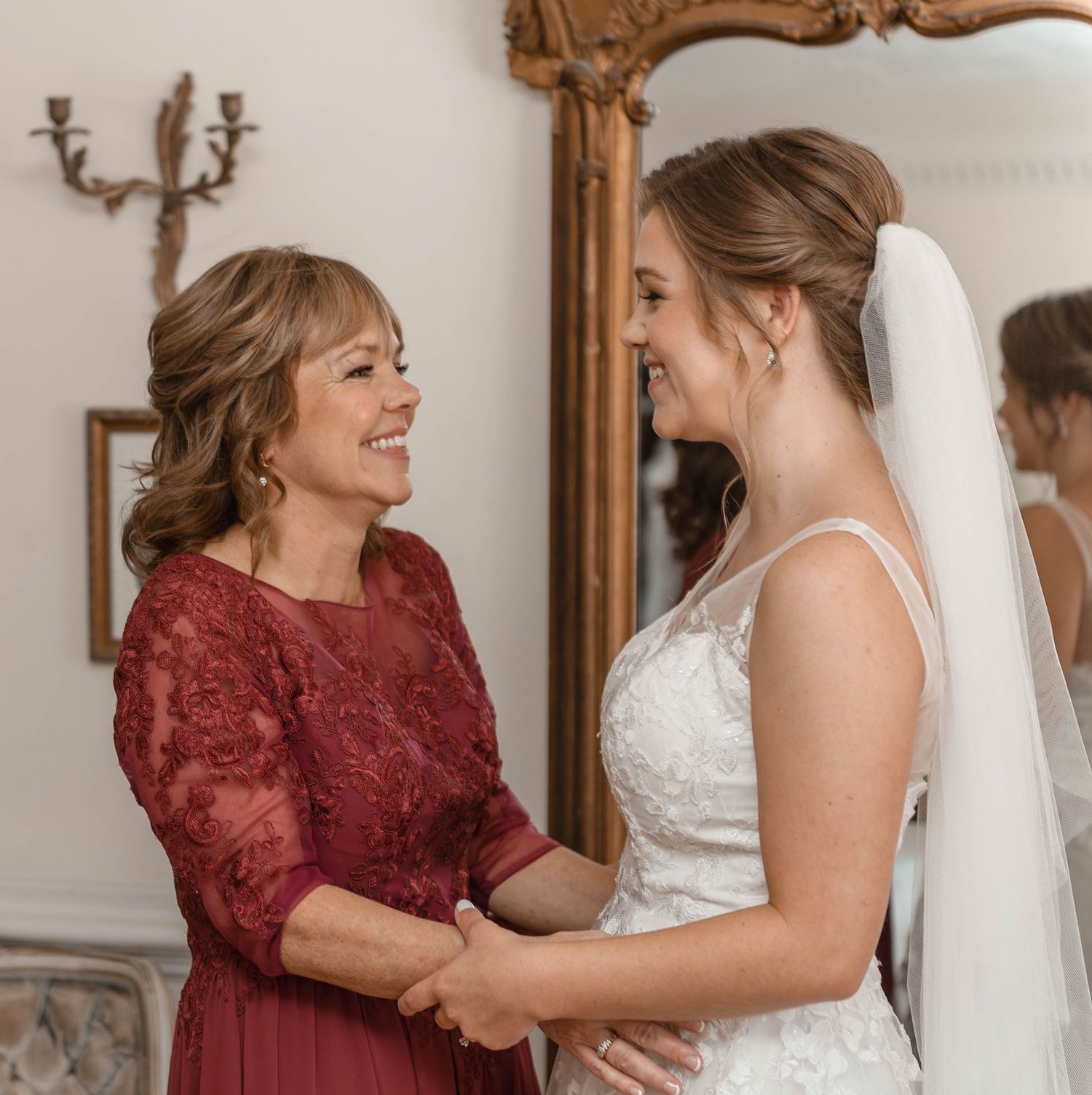 Bride in lace wedding gown shares a joyful moment with her mother in a burgundy lace dress.