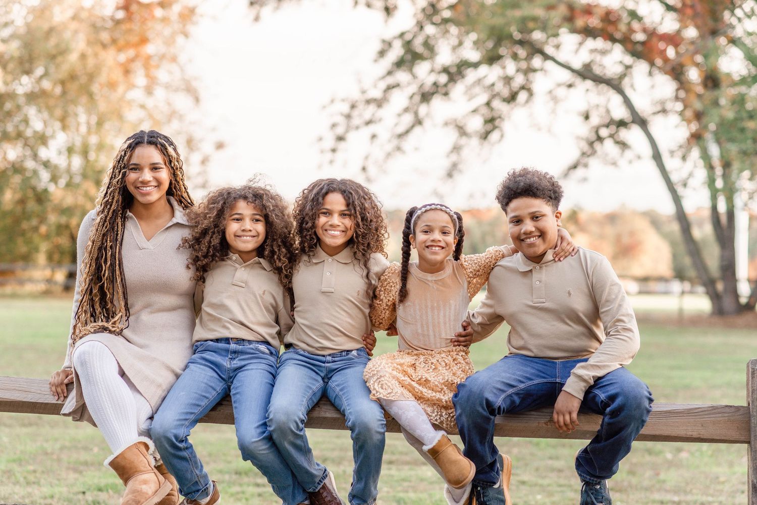 A mother and four children sit together on a wooden fence rail smiling in an autumn outdoor setting.