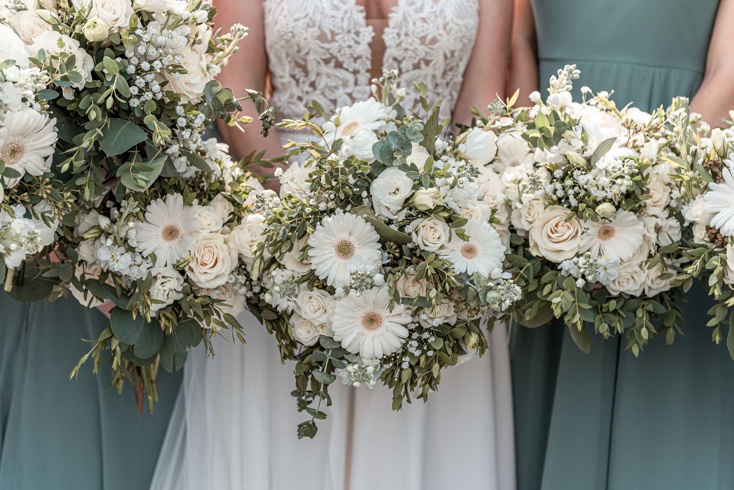 Bride and bridesmaids in sage green dresses holding matching white floral bouquets with greenery.