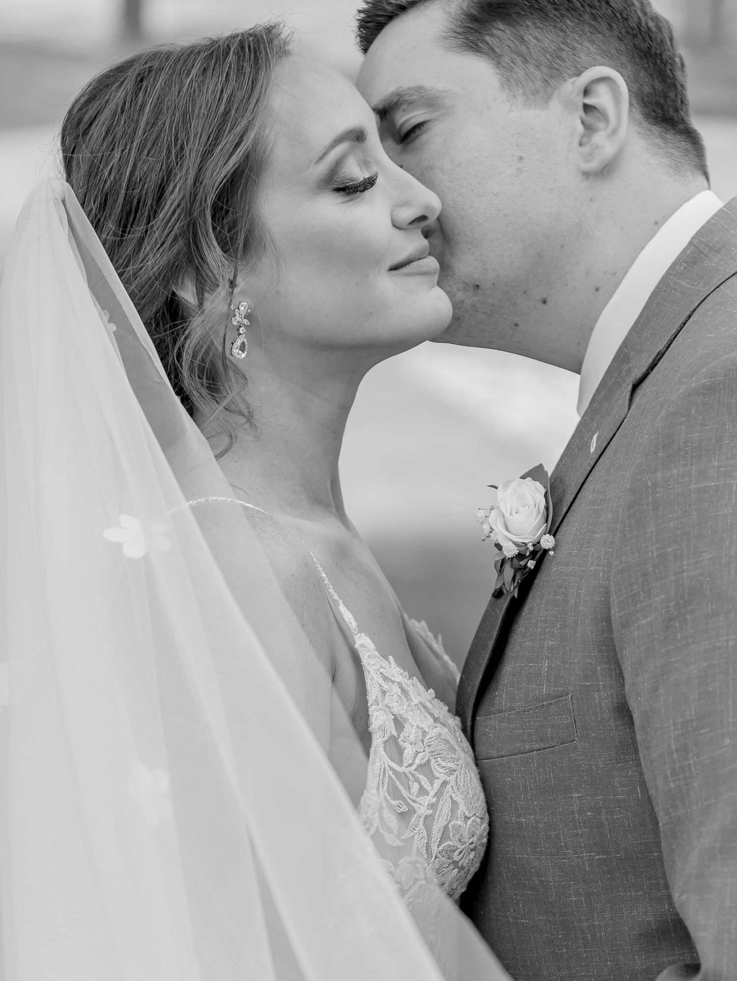 Black and white portrait of groom kissing bride on cheek as her veil flows around them.