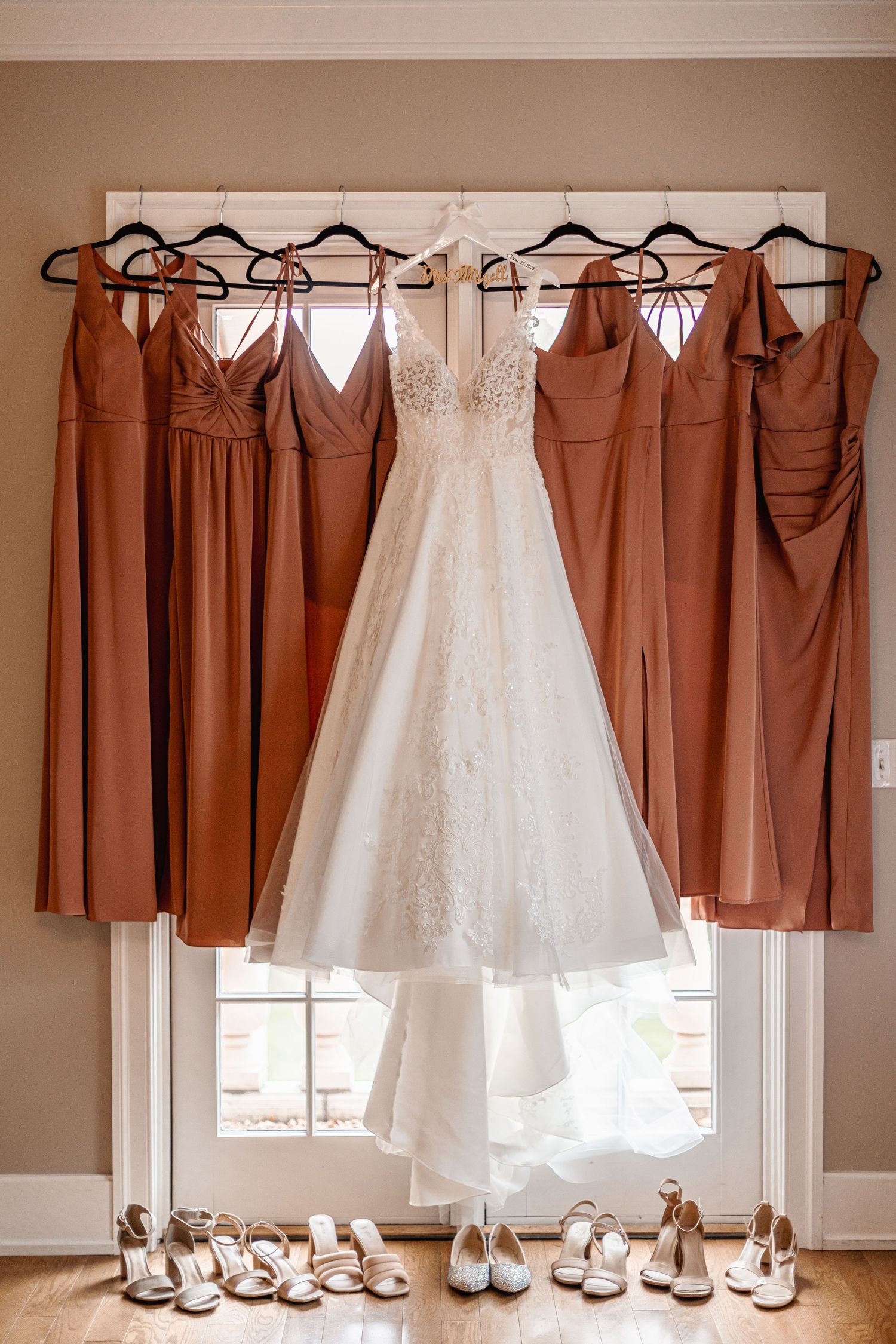White lace wedding gown flanked by rust-colored bridesmaid dresses hanging on a window with heels arranged below.