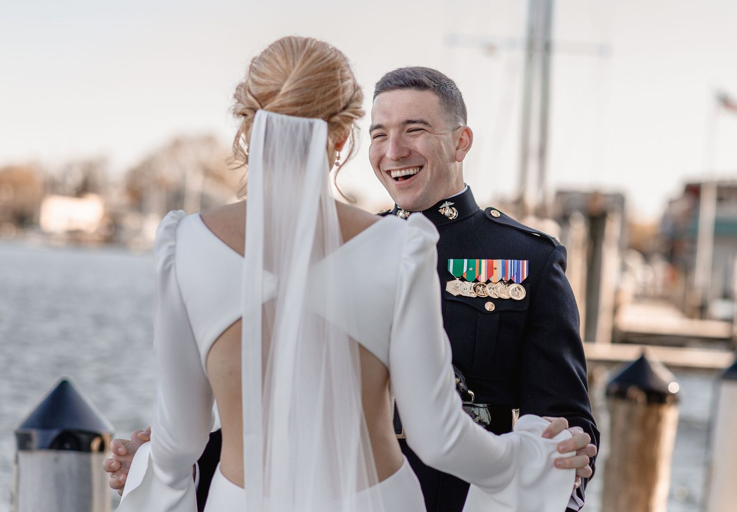 Marine groom in dress uniform laughs joyfully during first look with bride in white wedding gown on waterfront dock.