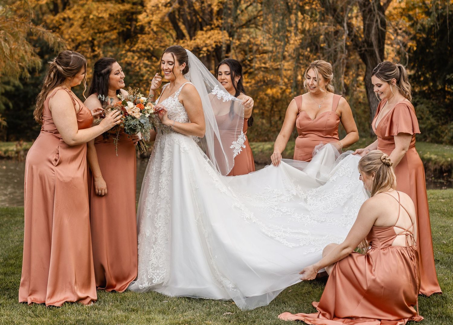 Bridesmaids in rust dresses laughing and fluffing bride's white lace gown during fall wedding photos.