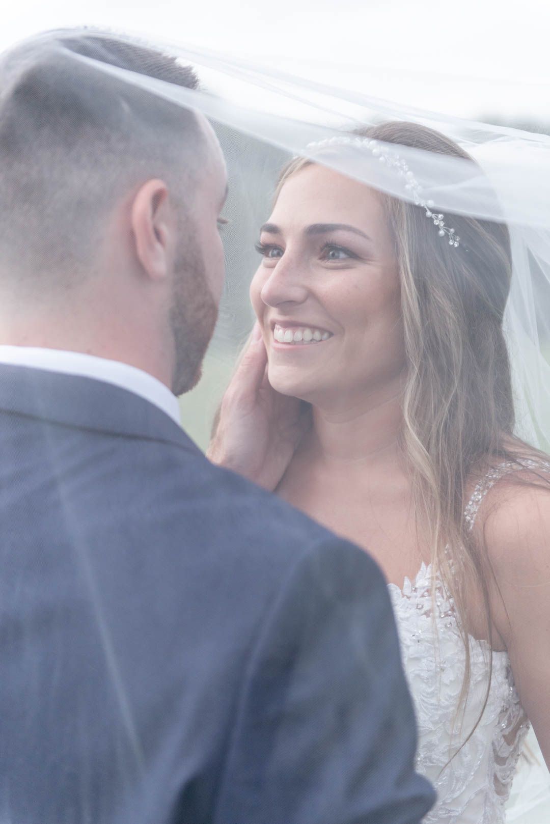 Smiling bride gazes at groom under a flowing veil, wearing lace wedding dress with hair accessories.