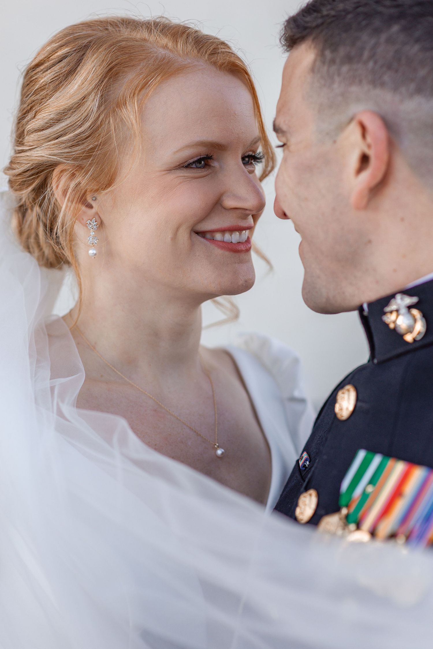 Radiant redhead bride smiles at Marine groom in dress uniform during intimate wedding portrait under veil.