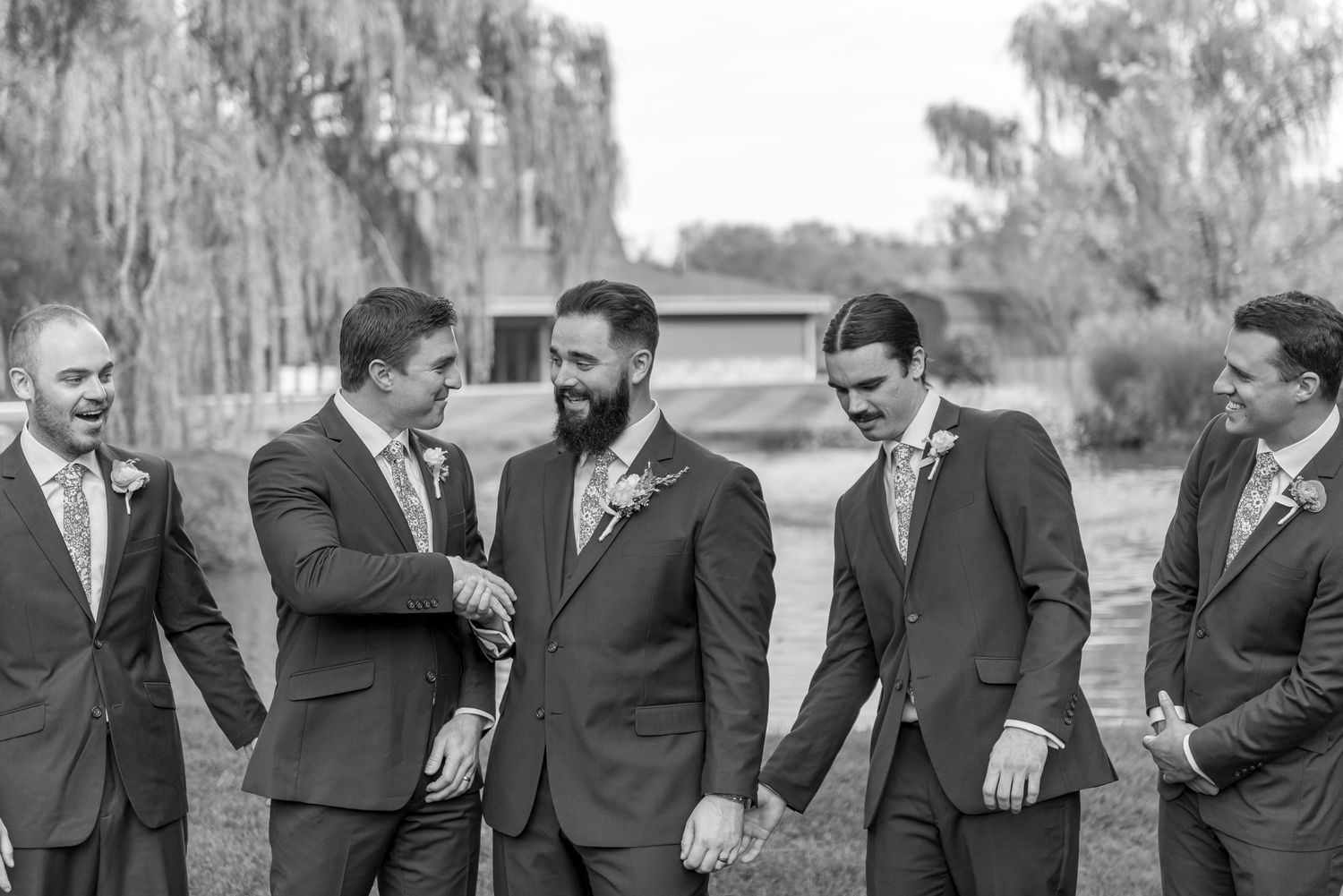 Groom and groomsmen in grey suits laughing together outdoors in black and white wedding photo.