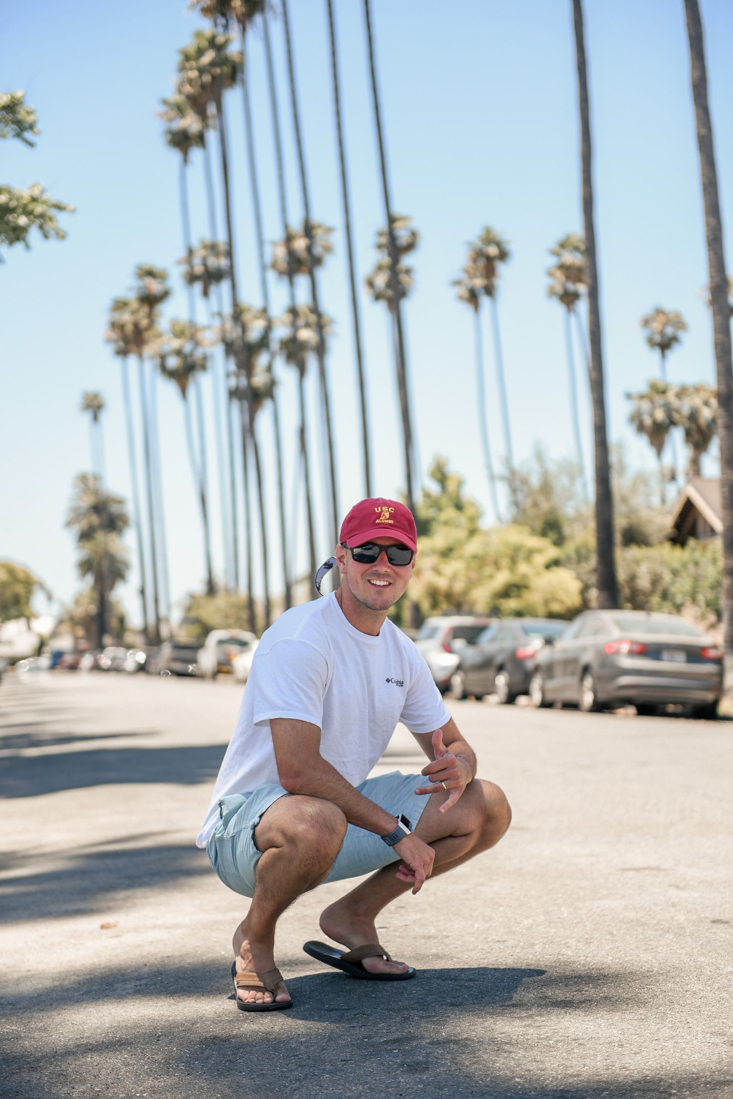 Man in white shirt and red cap crouches on street with tall palm trees in sunny California background.