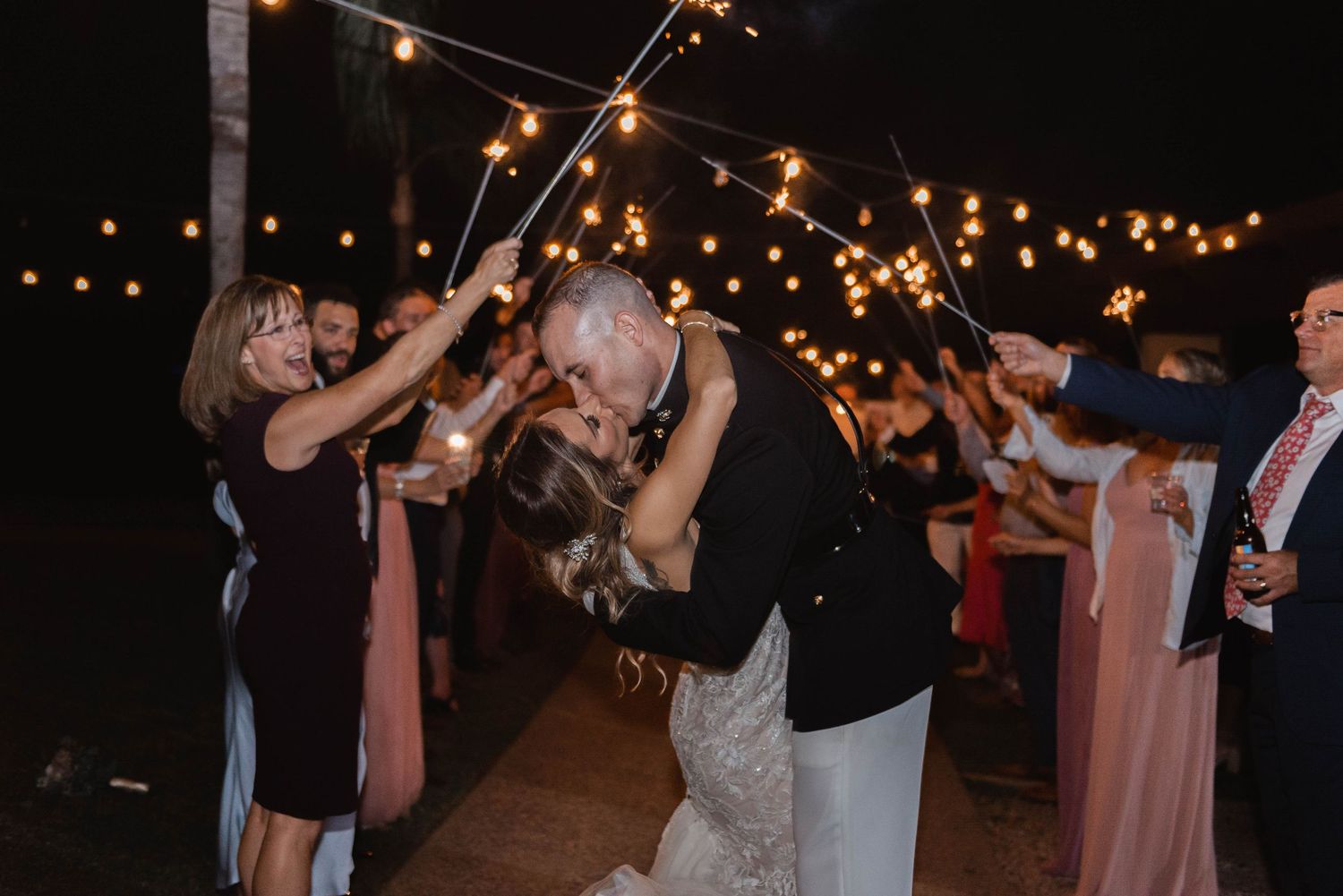 Couple sharing a romantic kiss during sparkler send-off at outdoor evening wedding reception.