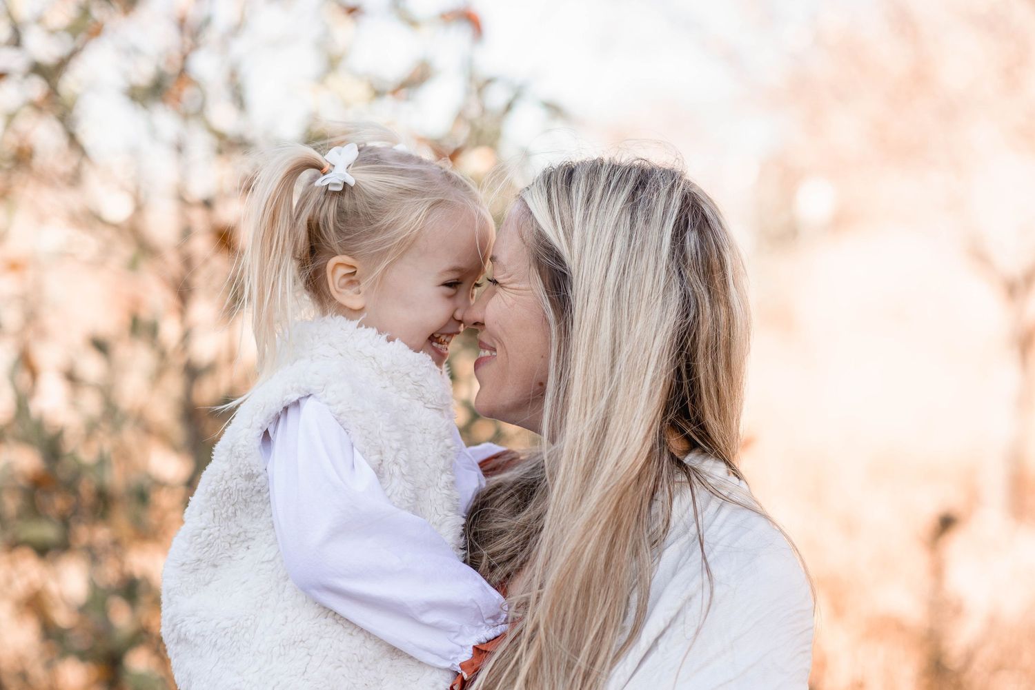 Mother and young daughter touching noses and smiling in warm autumn light during family portrait session.