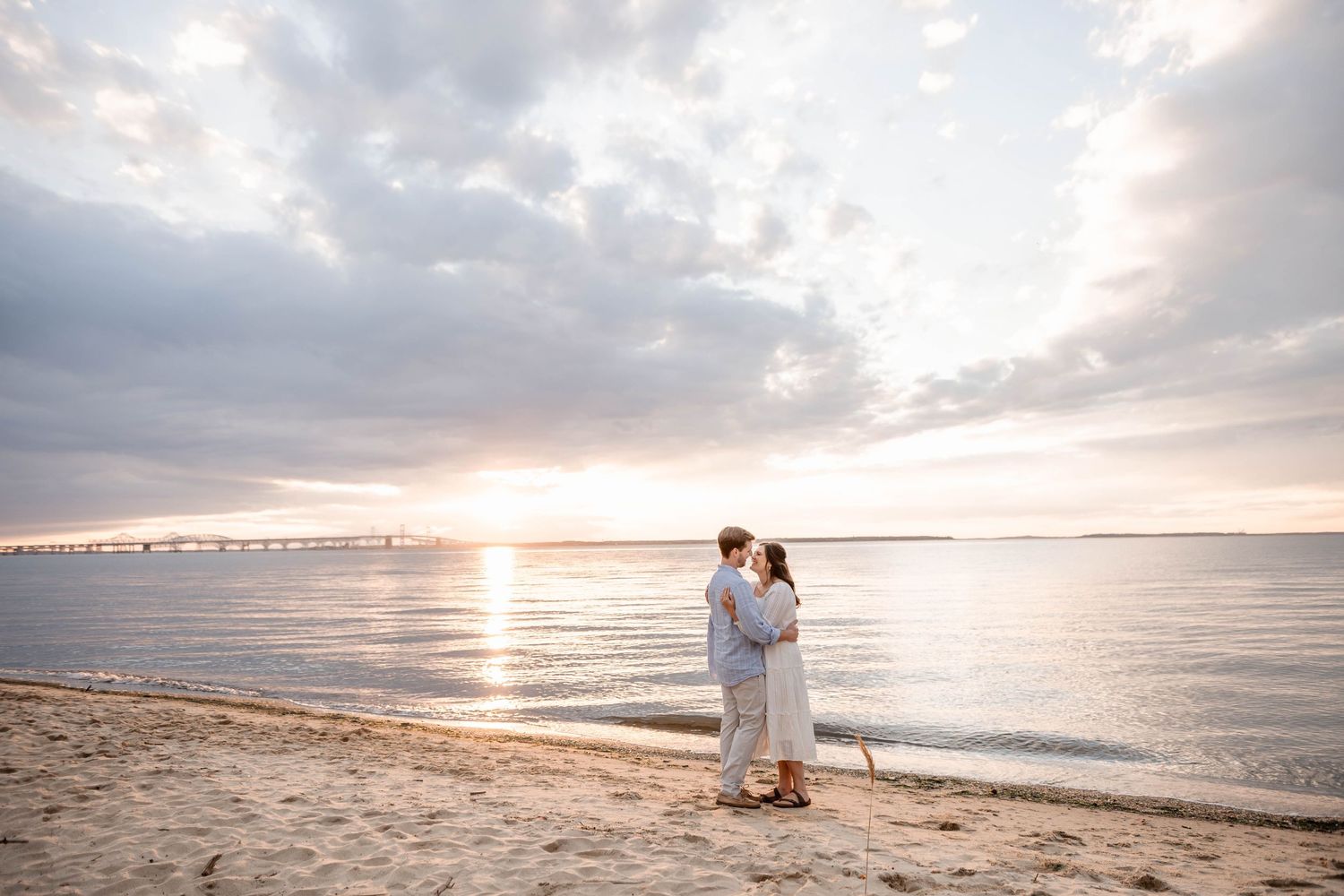 Couple embracing on sandy beach at sunset with dramatic cloudy sky and bridge in background.