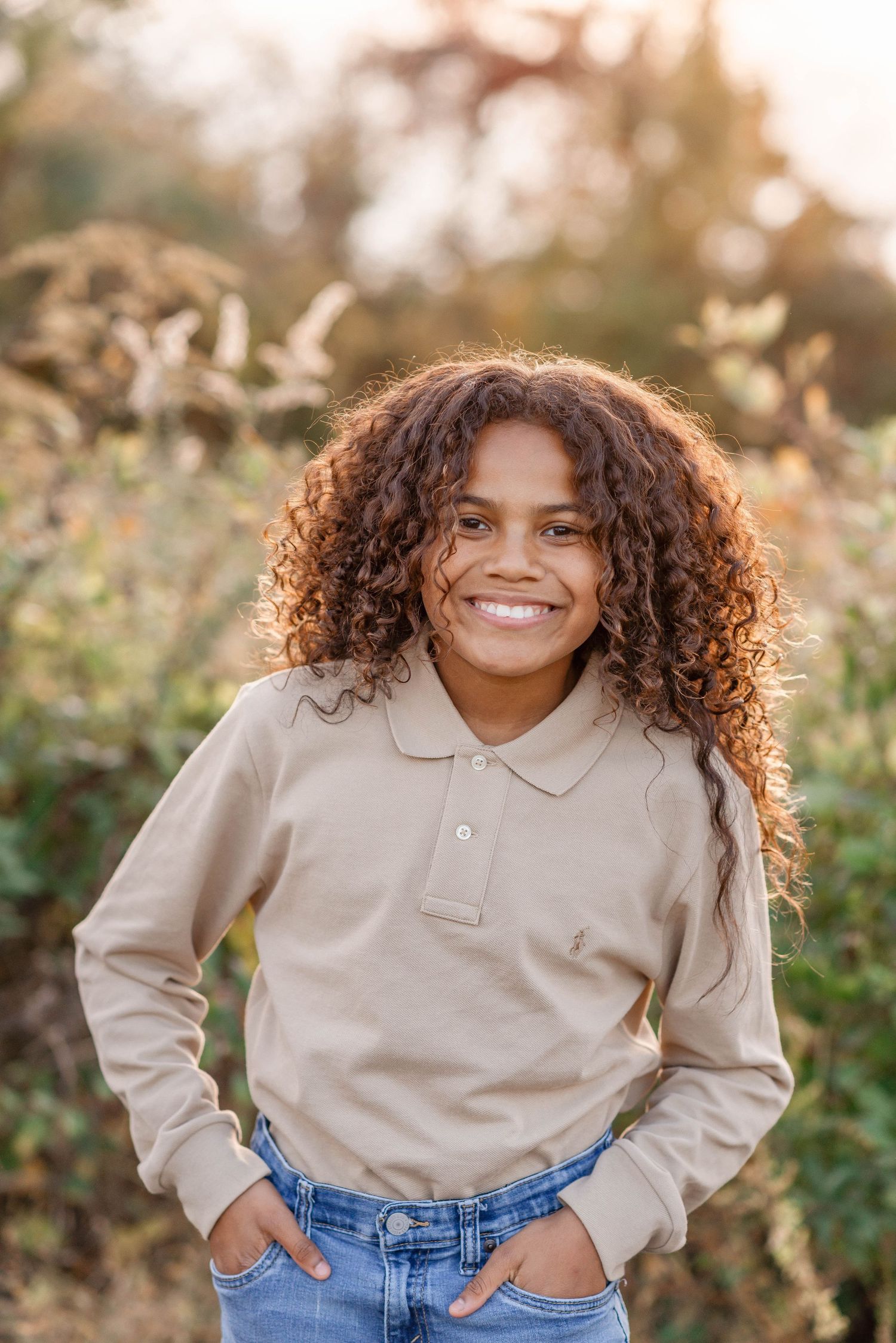 A smiling child with curly hair wearing a beige polo shirt standing outdoors in golden sunlight.