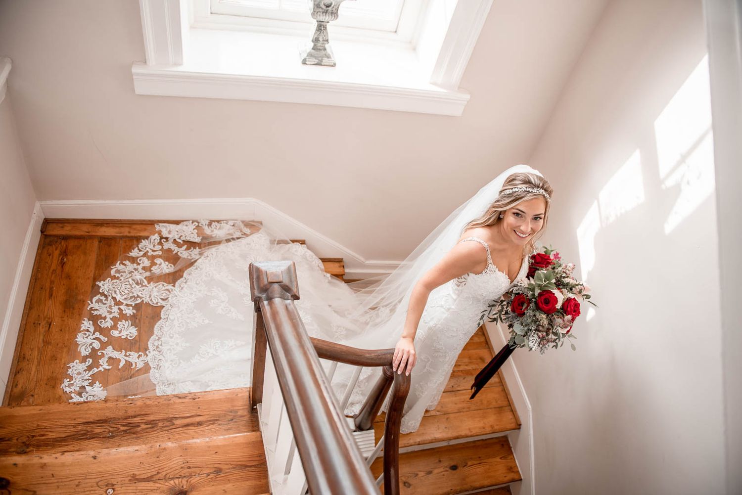 Bride in lace gown and veil descends a sunlit wooden staircase holding a red and white bridal bouquet.