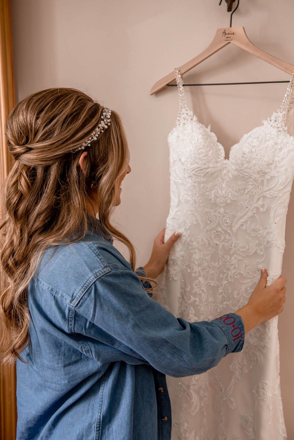 Bride with pearl headpiece admires her lace wedding dress on a hanger before the ceremony.