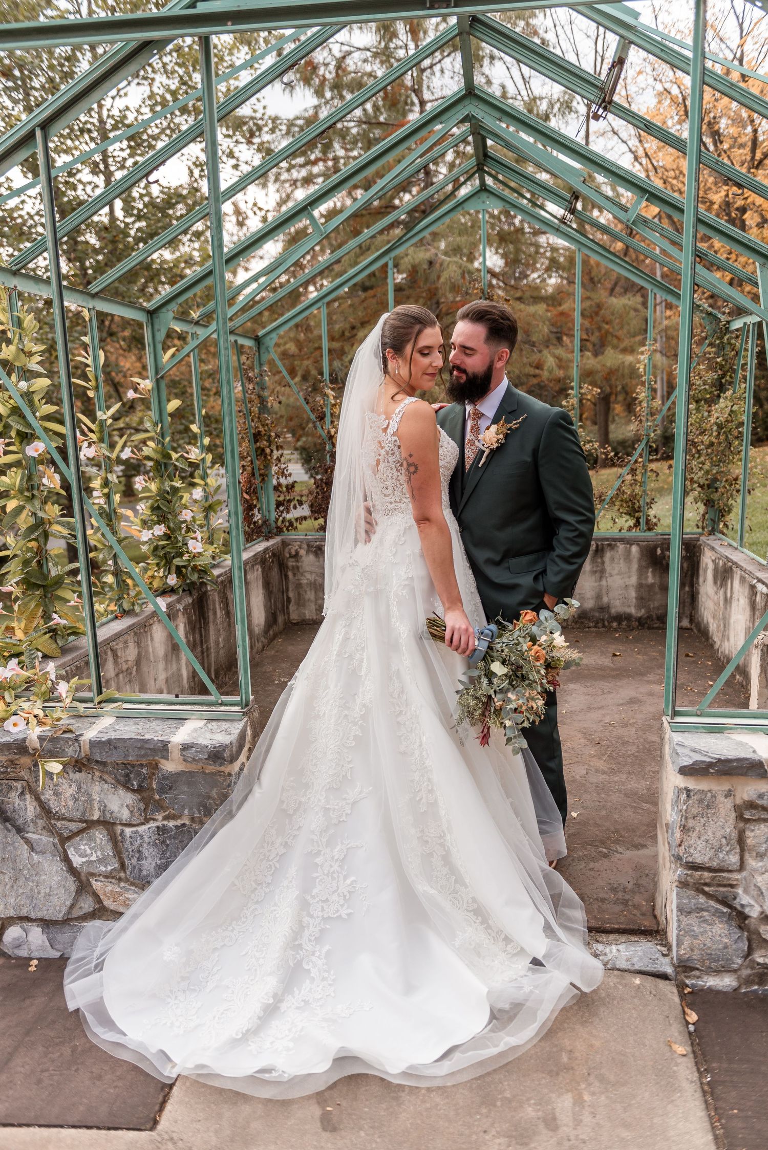 Bride and groom posing inside a glass greenhouse, she in lace gown with veil, he in dark green suit.