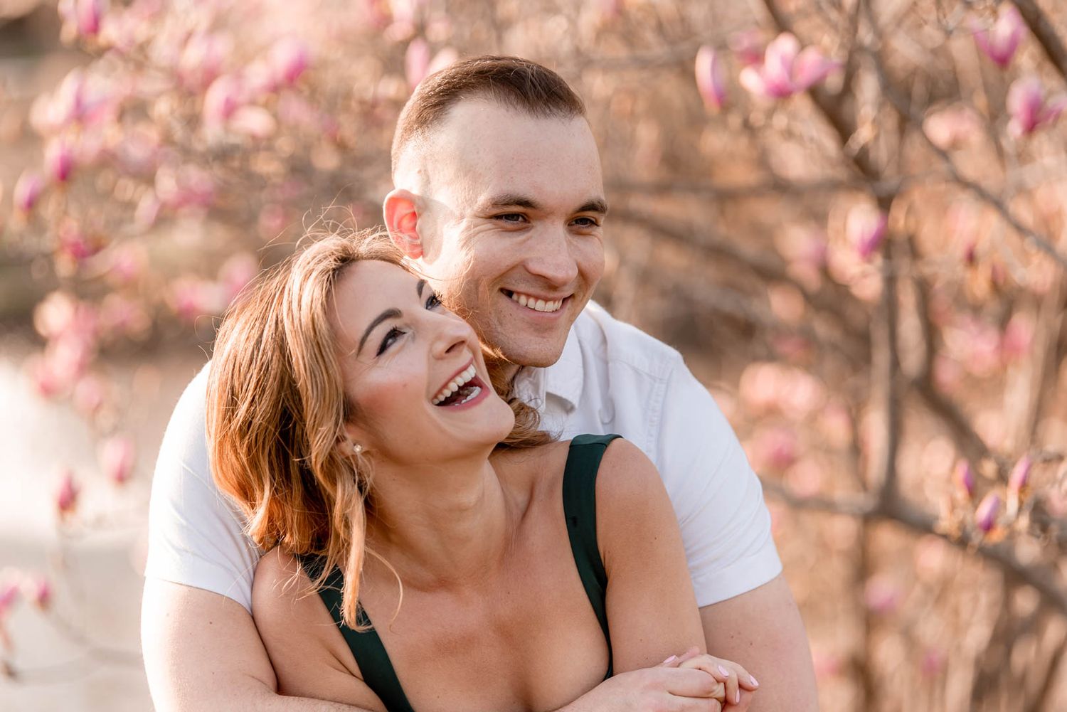 Joyful couple laughing and embracing in front of blooming pink magnolia trees during engagement photos.