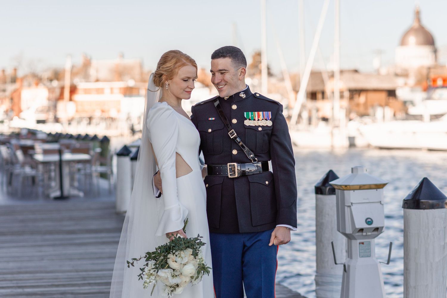 Smiling couple in wedding attire stand together on a dock with a scenic harbor backdrop.