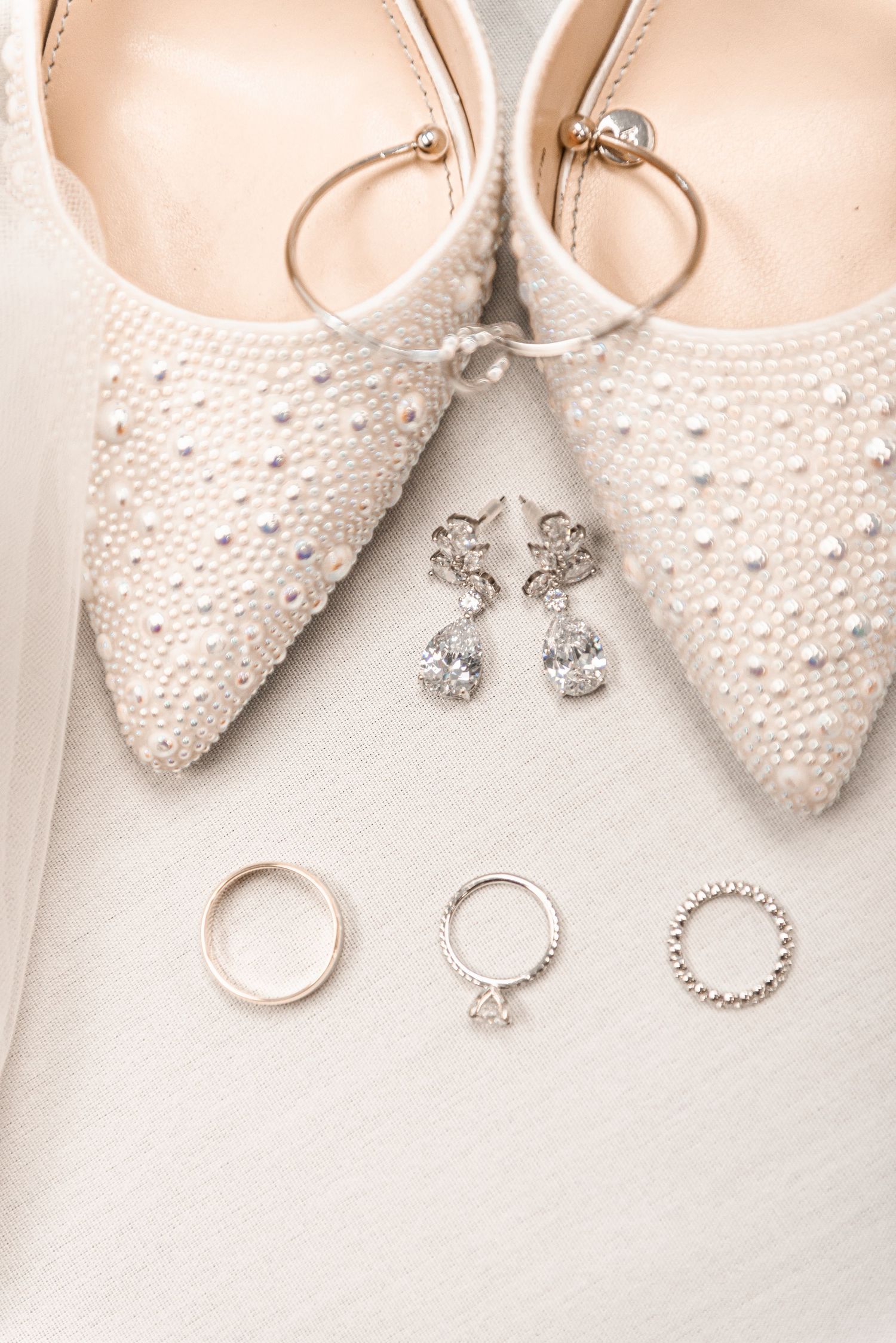 Pearl-studded bridal heels displayed with crystal drop earrings and rose gold wedding bands on a soft white background.